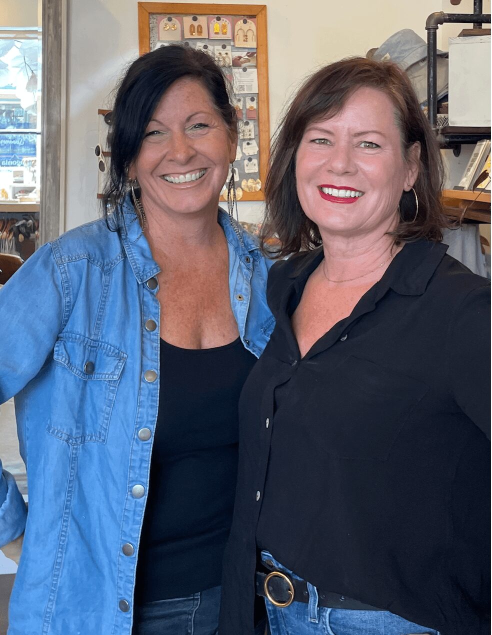 Two women smiling indoors, one wearing a denim jacket and the other a black shirt, with shelves and a window in the background.