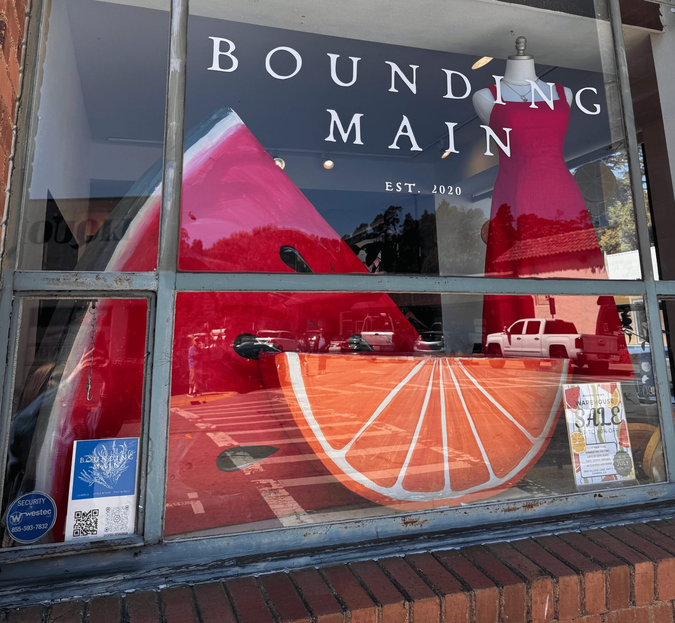 Store window display with large pink and red dresses, text 'Bounding Main' and 'Est. 2020', and a large orange slice graphic.