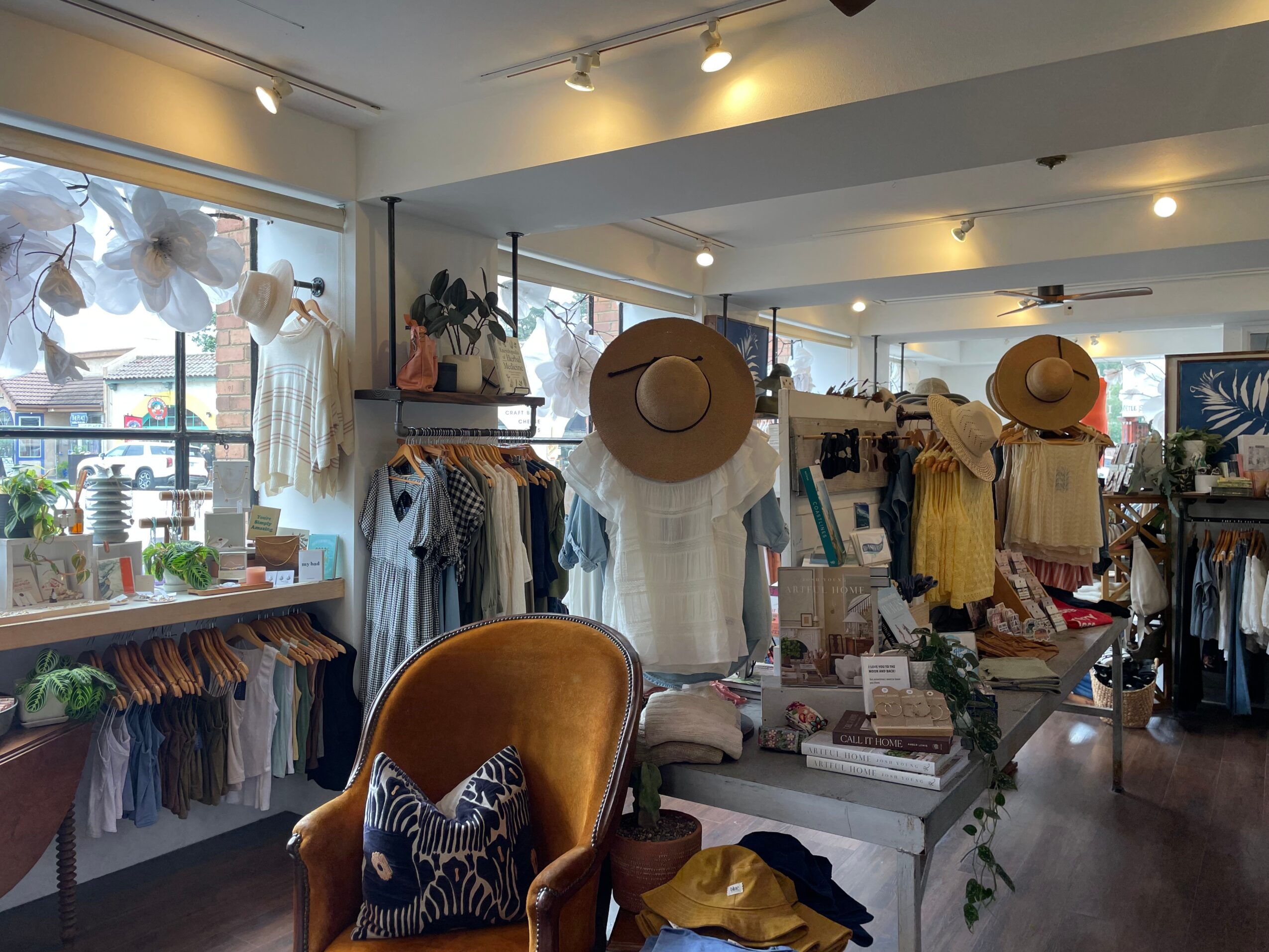 Clothing store interior with racks of clothes, hats, and a seating area, bright natural light from large windows, and decorative items.