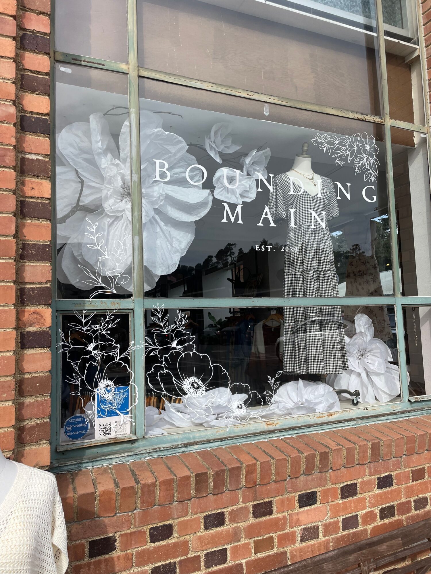 Broken glass window with floral decorations and a person in the reflection, brick wall below, and a sign reading 'BOSCO MAINE'.