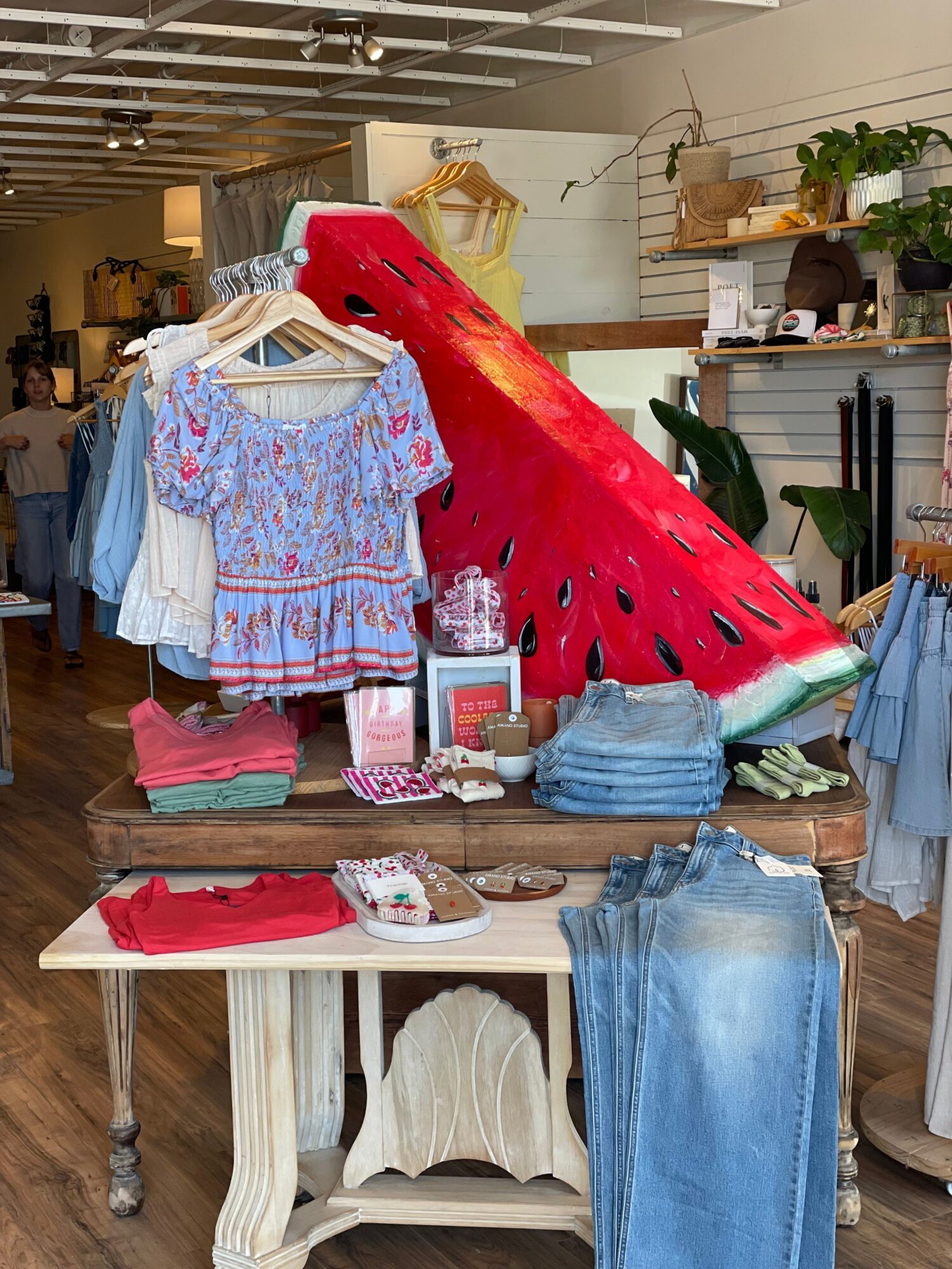 Display of clothing and accessories on a wooden table in a store, with a watermelon-shaped decoration in the background.