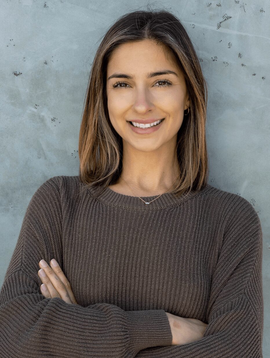 Young woman with shoulder-length brown hair smiling, wearing a dark brown sweater, standing against a gray wall.