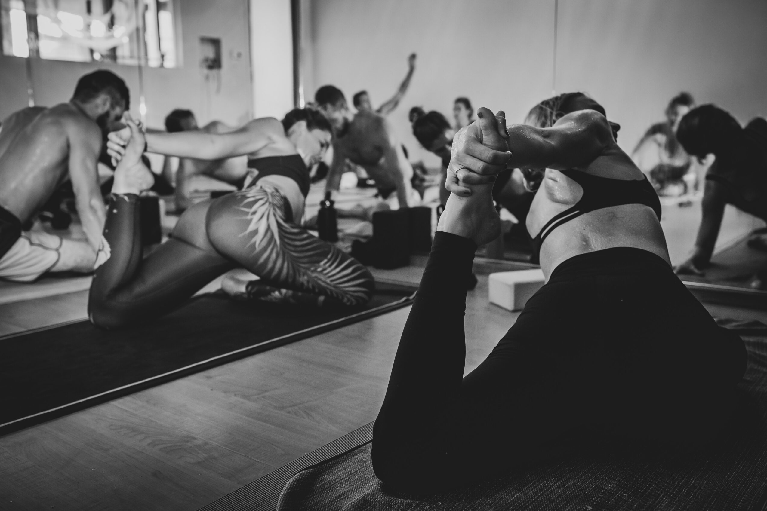 Group of people practicing yoga in a studio, performing various poses on mats, with a person in the foreground holding a pose.