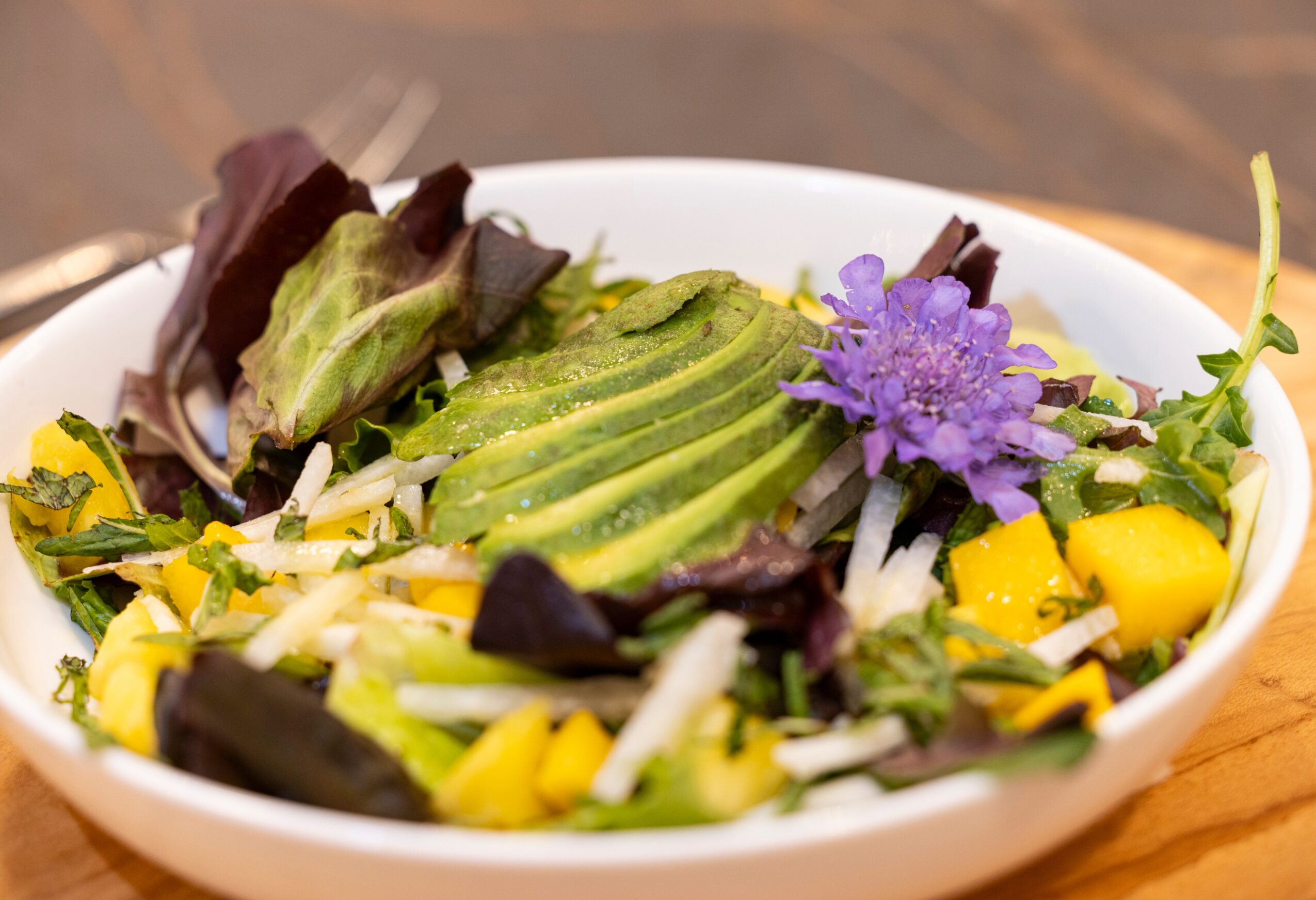 Fresh salad with sliced avocado, purple flower, and mixed greens in a white bowl on a wooden surface.