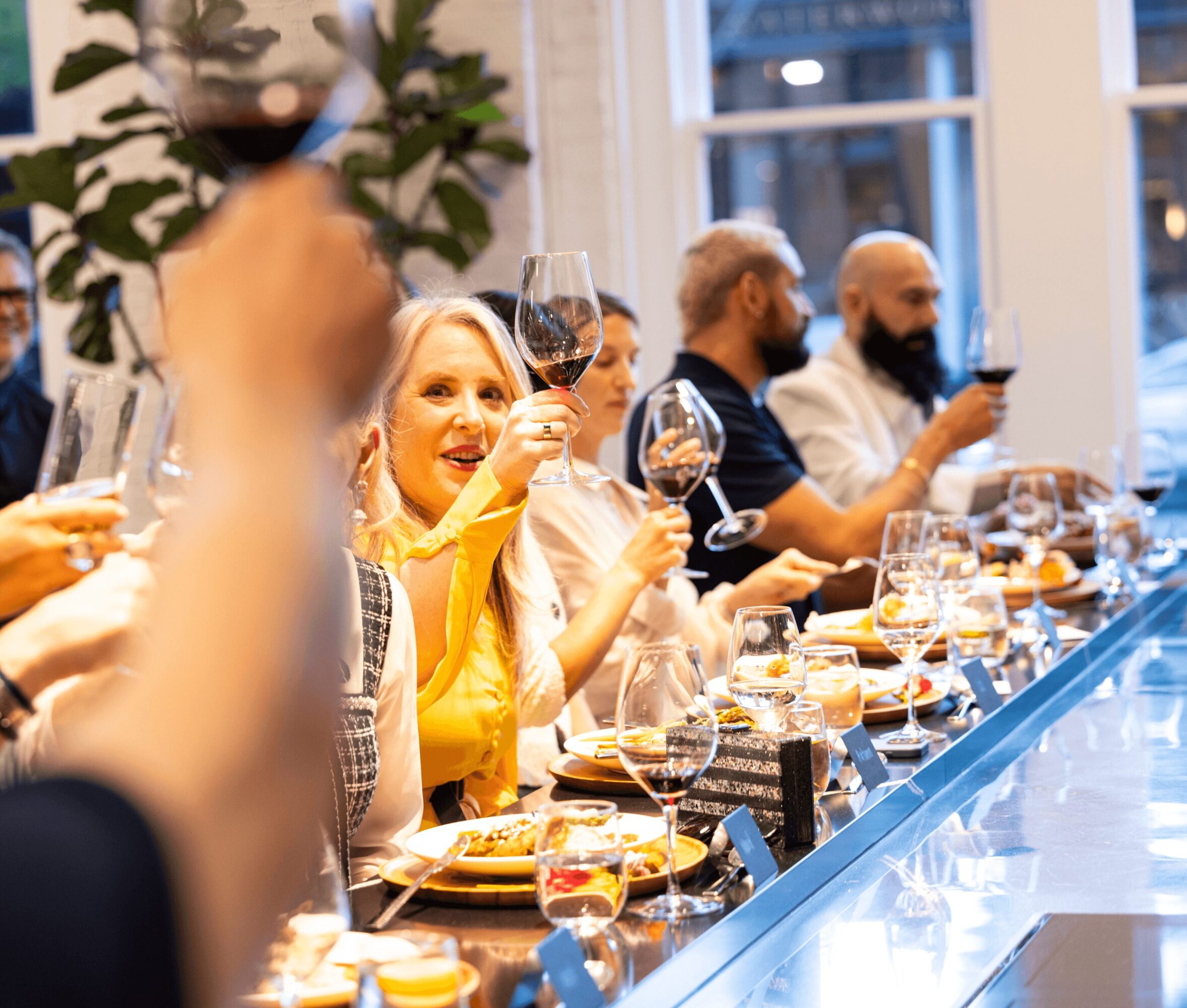 People sitting at a dinner table raising glasses in a toast, with food and drinks on the table, in a well-lit room.