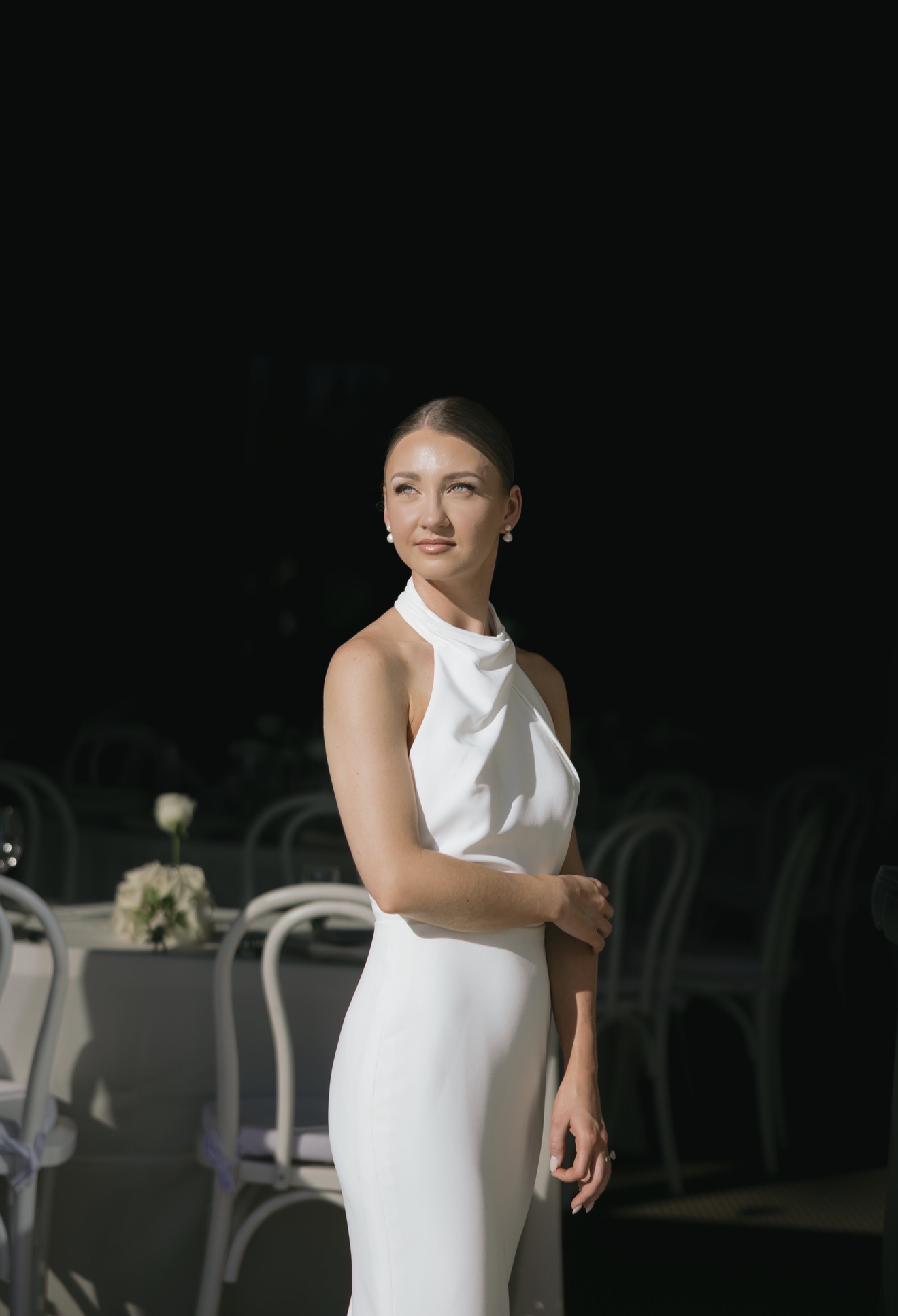 Woman in white dress standing outdoors at night, with tables and chairs in the background.