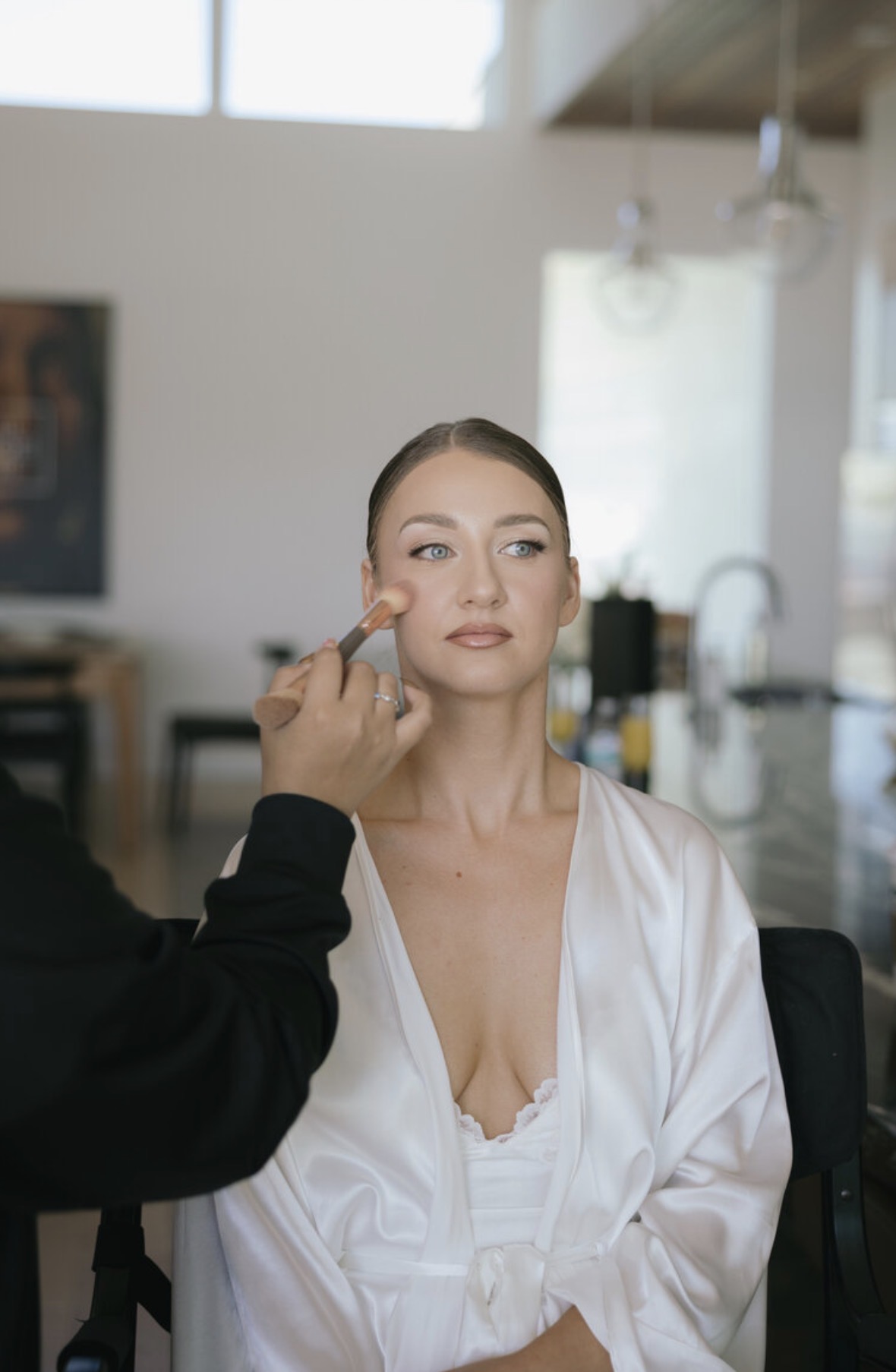 Woman with makeup being applied, sitting in a bright room with large windows and hanging lights.