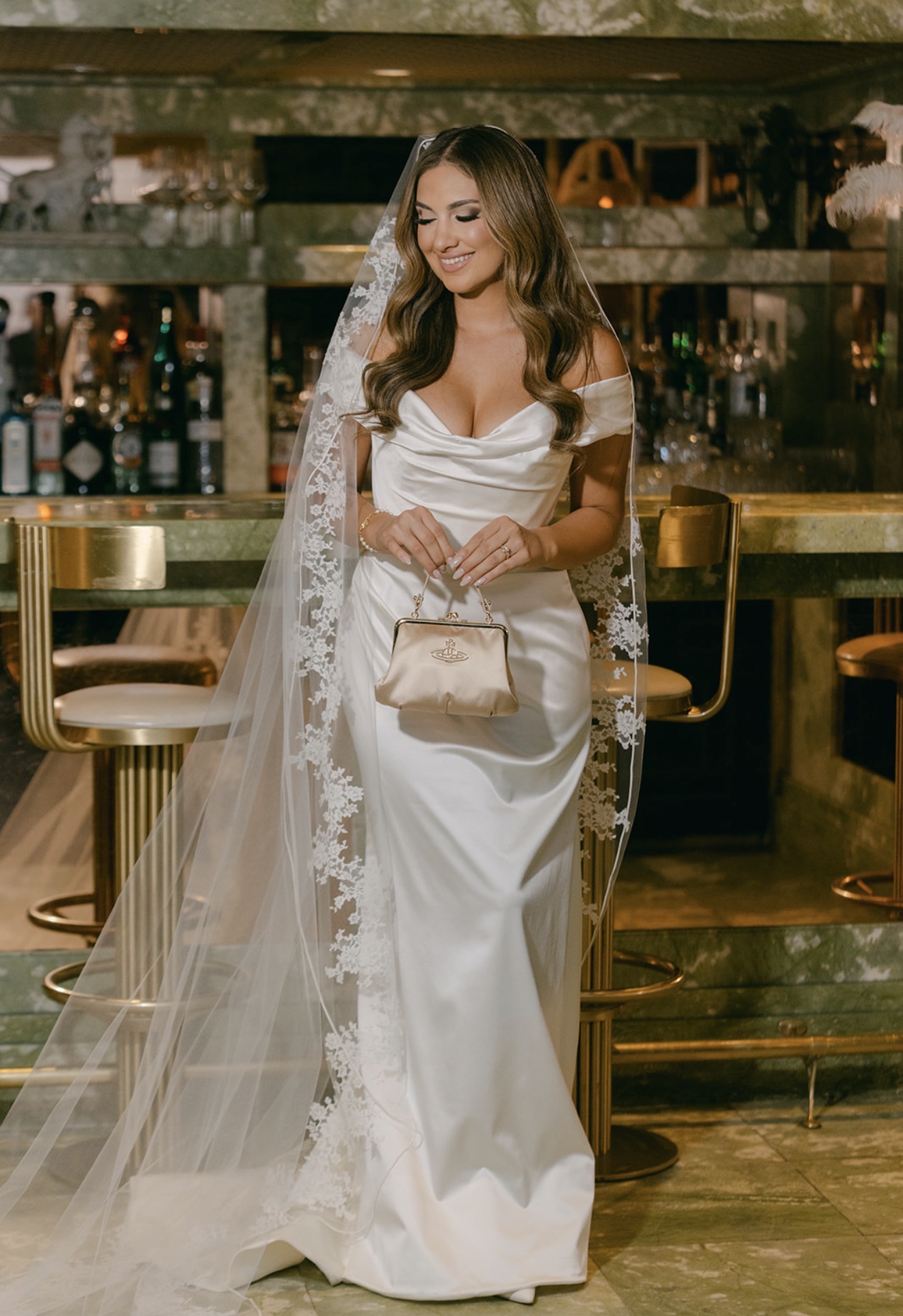 Woman in wedding dress with veil holding a clutch, standing in a bar or restaurant setting.