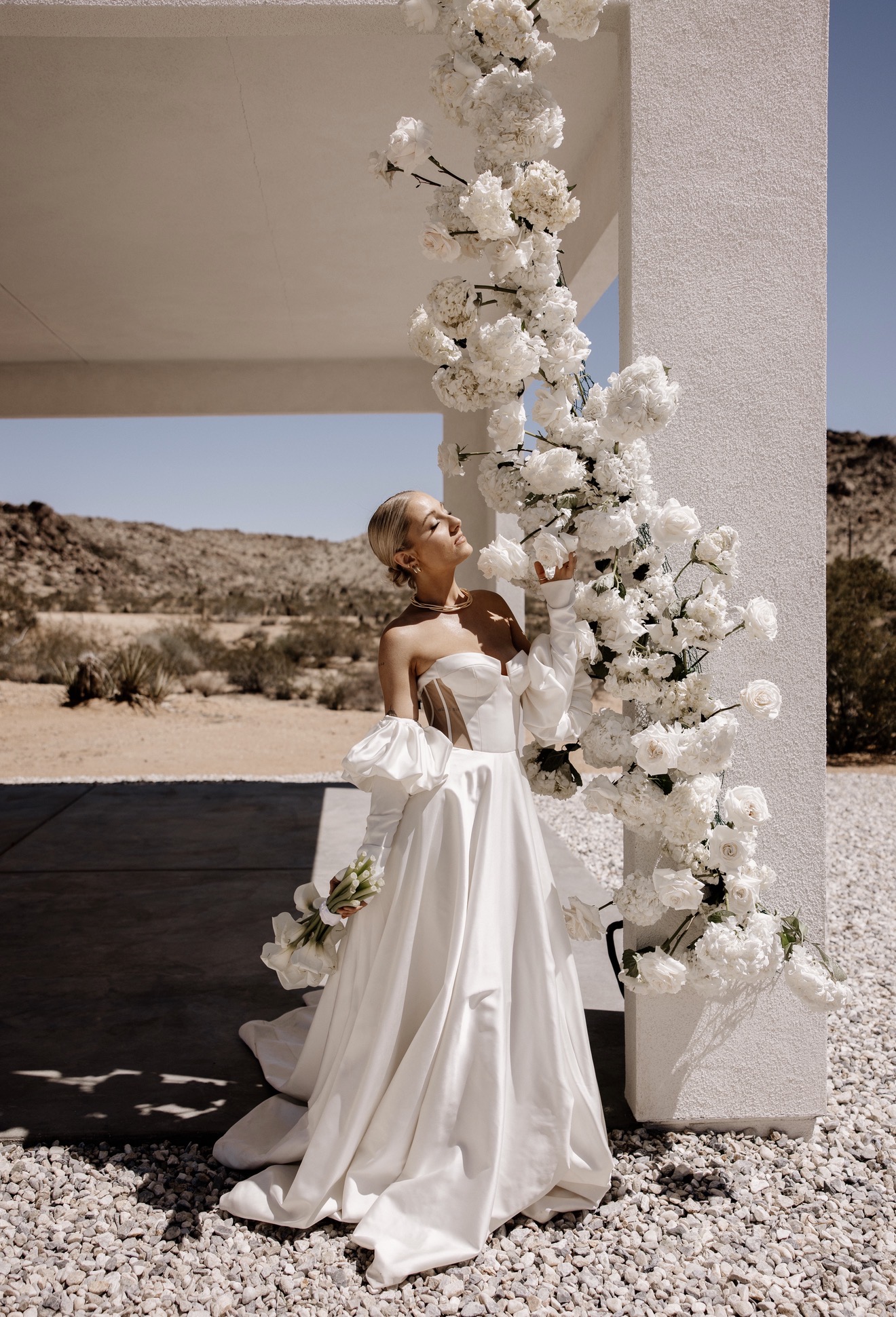 Woman in a white dress standing next to a tall floral arrangement outdoors, desert landscape in background.