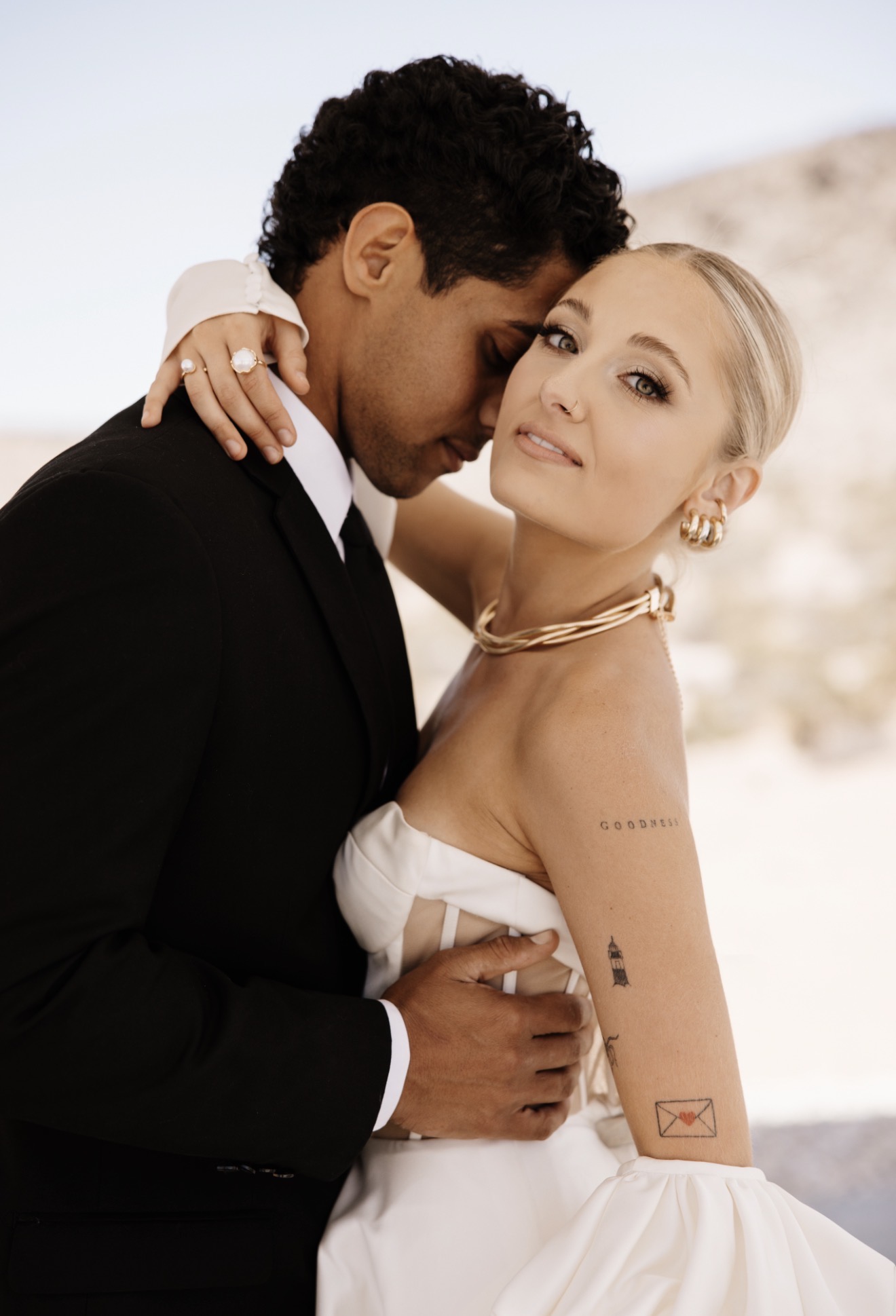 A couple embraces outdoors, with the man in a black suit and the woman in a white dress, both smiling gently.