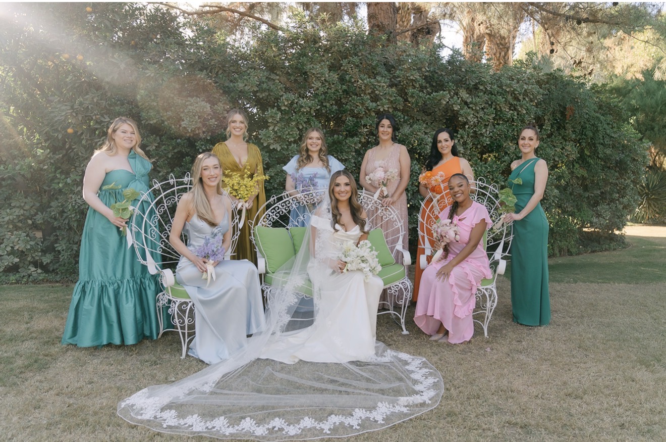 Group of nine women, one in wedding dress, outdoors with greenery, some seated and some standing, sunlight shining through trees.