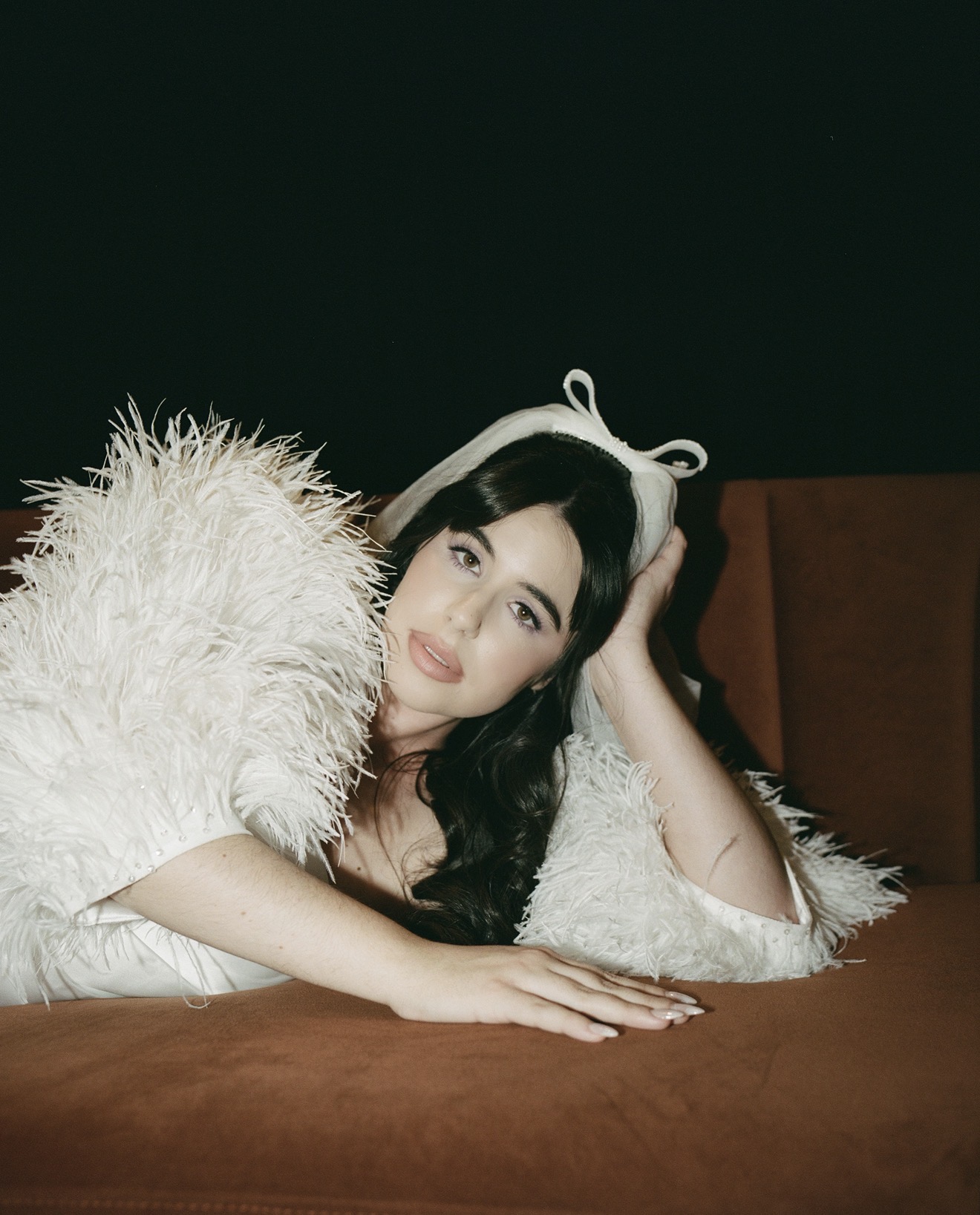 Woman lying on a brown surface, wearing a white furry garment and a headband with bunny ears, resting her head on her hand.
