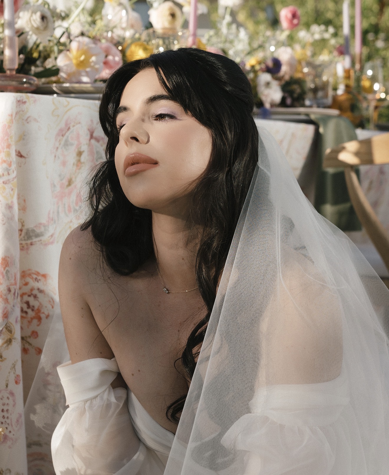 Young woman with dark hair and a veil, sitting outdoors with floral decorations in the background.