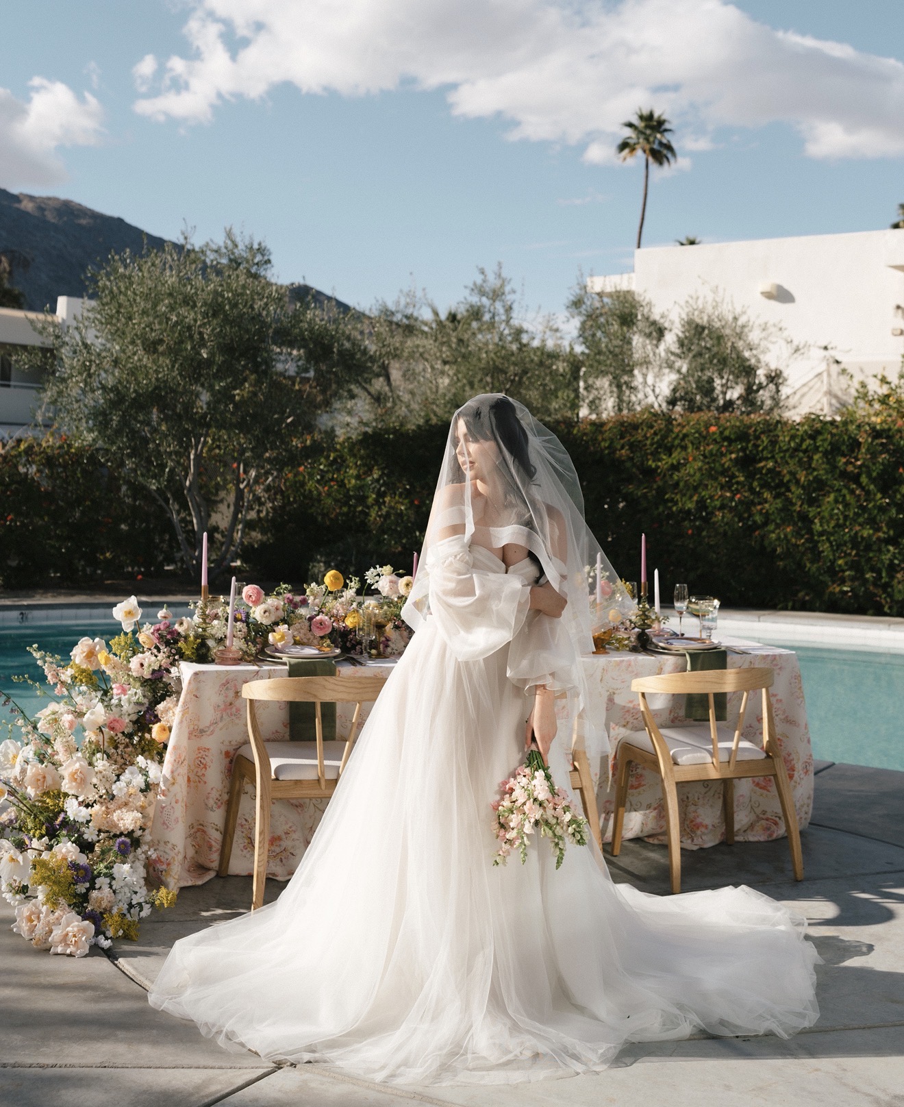 Bride in wedding dress holding bouquet, standing outdoors near decorated table with flowers, trees, and a pool, sunny sky.