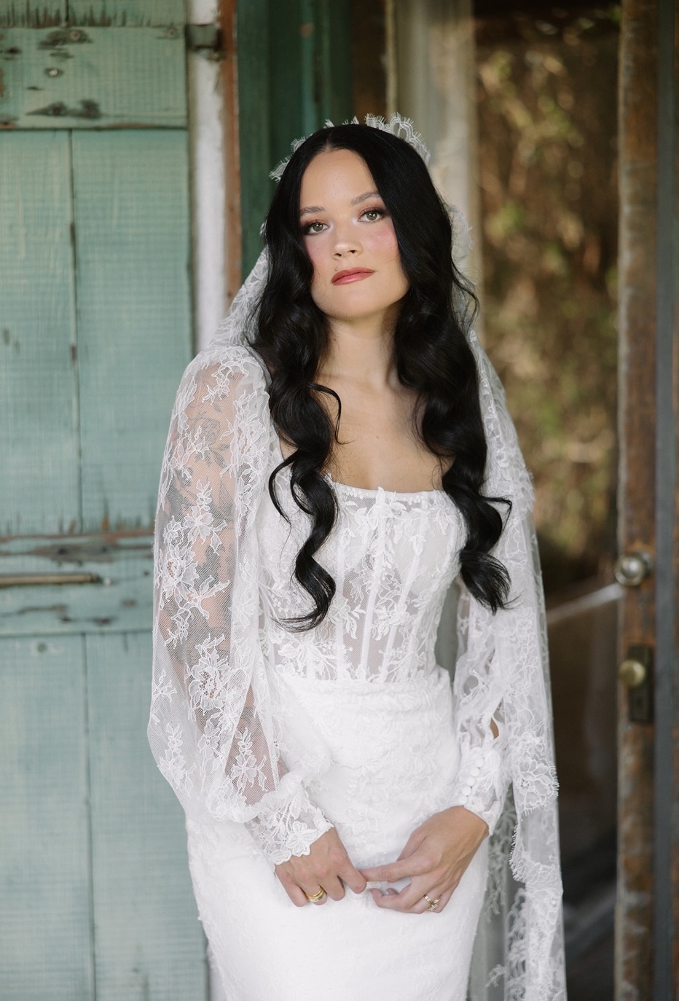 Woman with long dark hair in a white lace dress standing outdoors near a weathered green door.