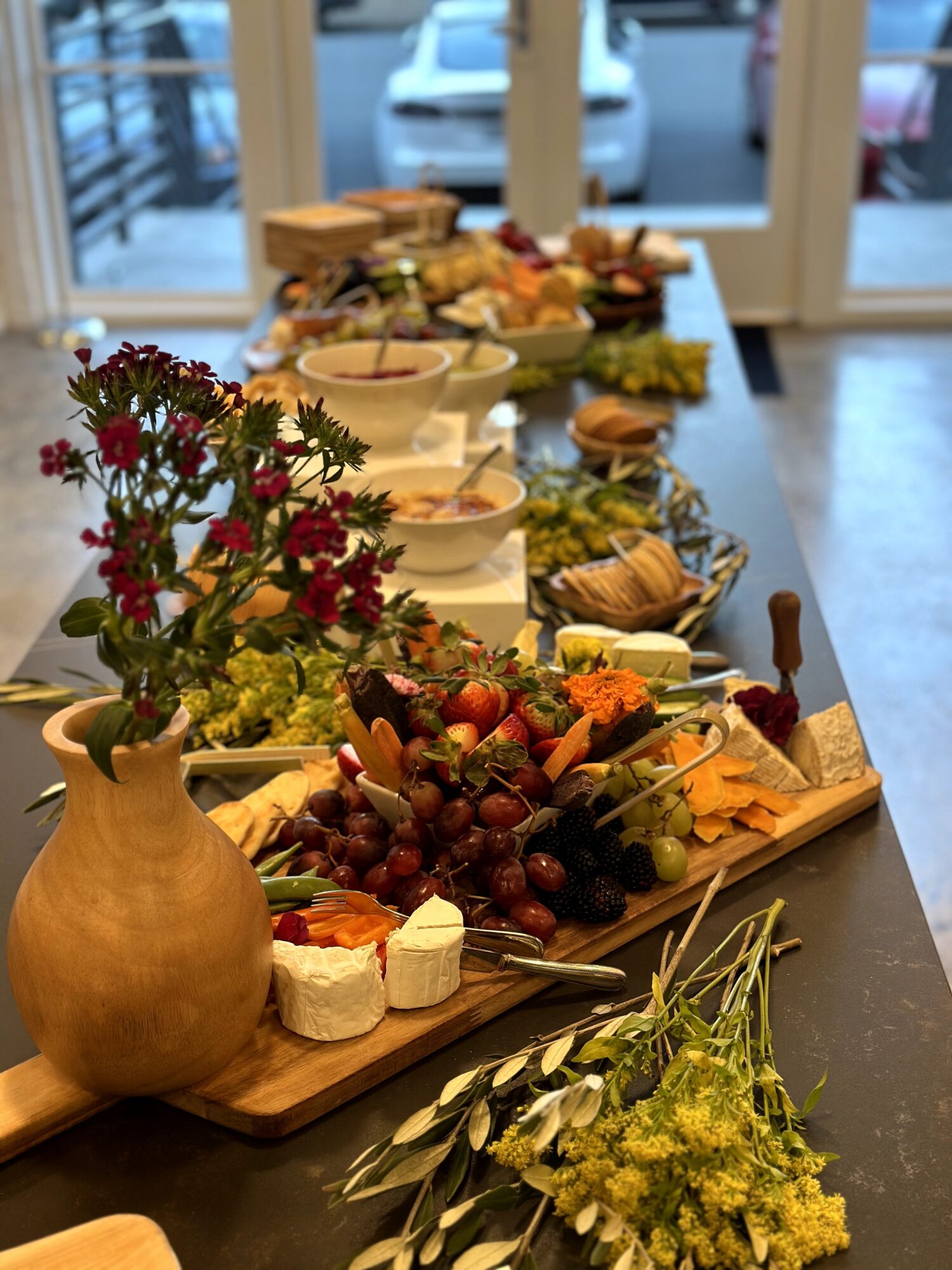 Table with various foods and a vase with red and green flowers, set indoors near glass doors.