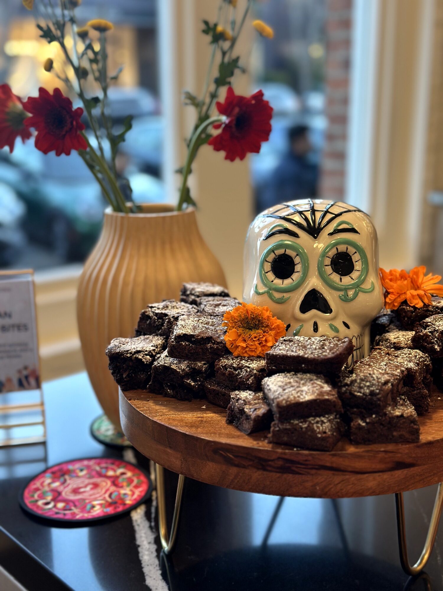Decorative sugar skull surrounded by chocolate treats with orange flowers, in front of a vase with red and yellow flowers, near a window.