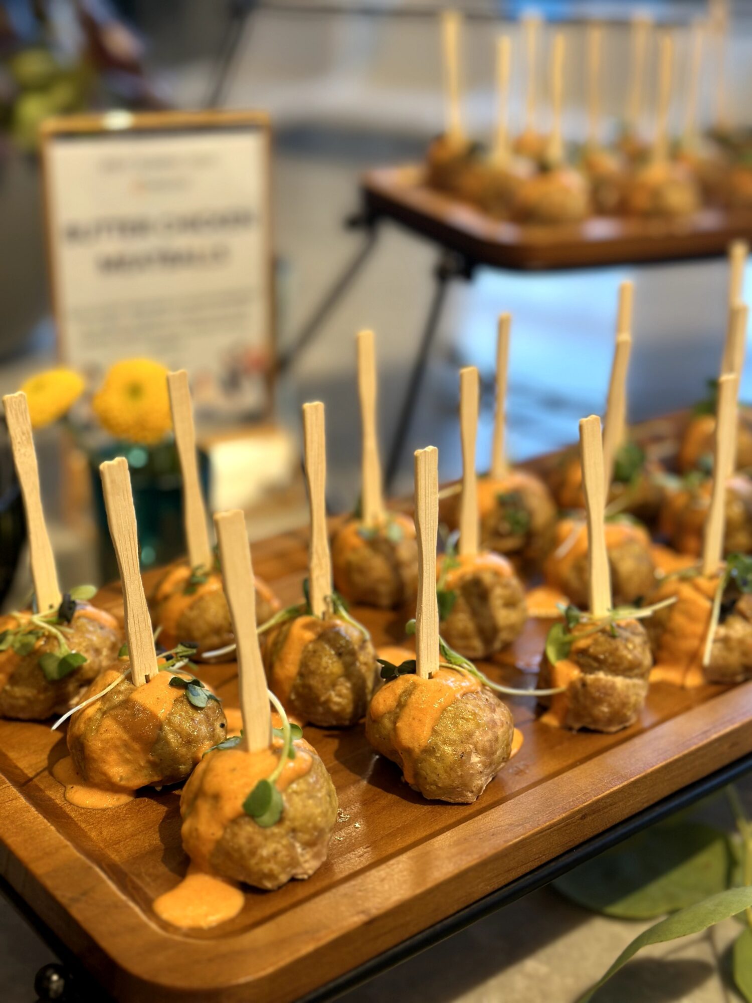 Close-up of skewered appetizers on a wooden tray, with a blurred background of a sign and more skewers.