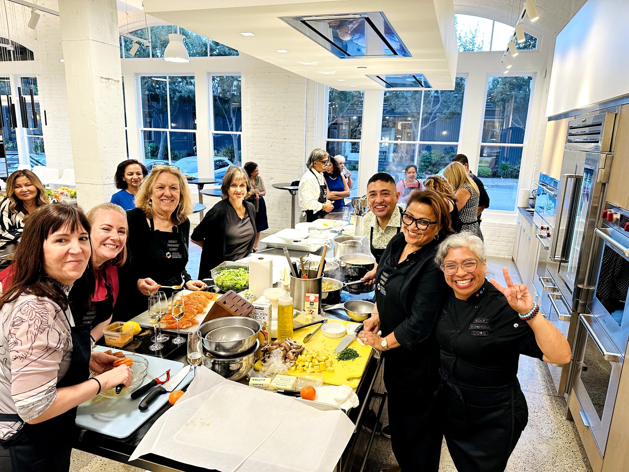 Group of people gathered around a kitchen counter preparing food, smiling, with large windows and modern decor in the background.