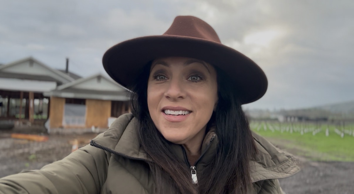 Woman outdoors wearing a wide-brimmed hat and jacket, smiling, with cloudy sky and buildings in background.