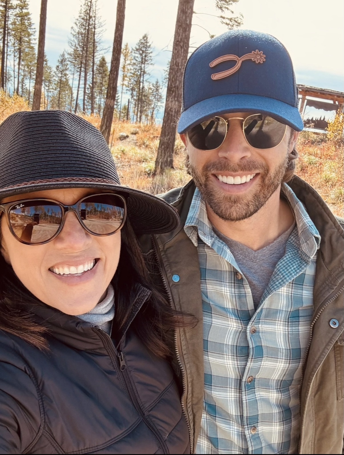 Two smiling people outdoors, one wearing a black hat and sunglasses, the other wearing a blue cap and sunglasses, with trees in the background.