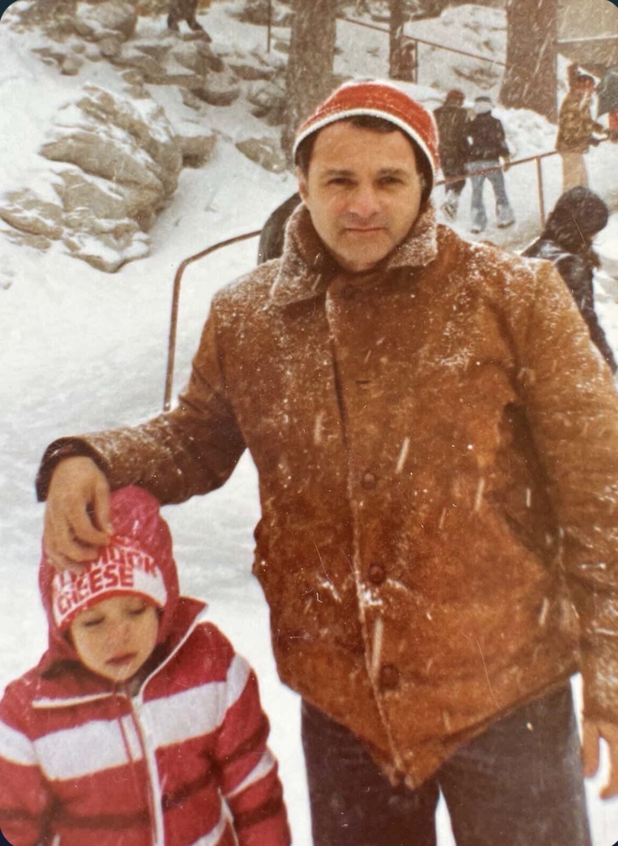 Man in brown jacket and red hat standing in snowy landscape with children and trees in background.