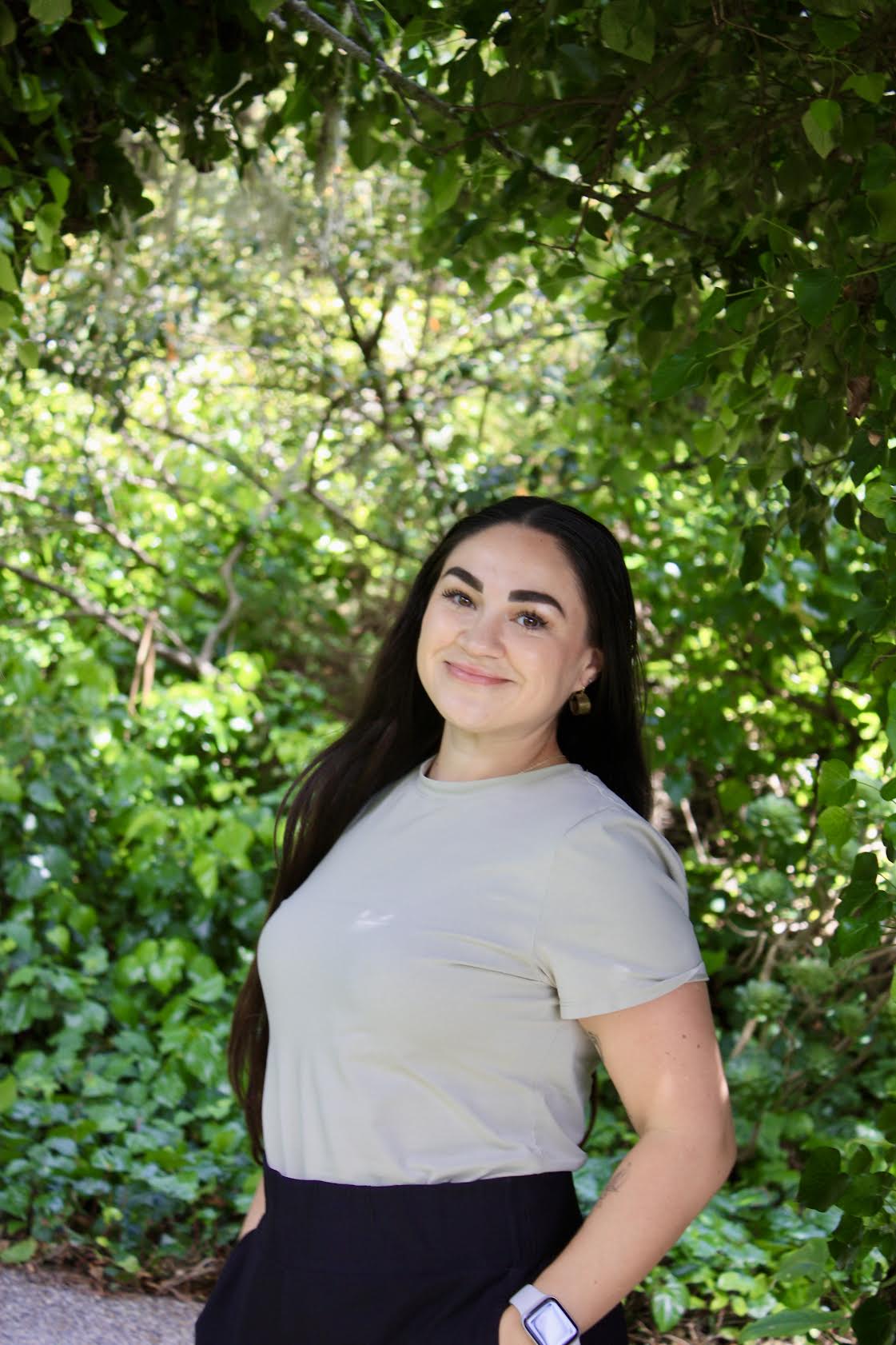 Young woman with long dark hair smiling outdoors surrounded by green foliage, wearing a light-colored t-shirt and dark pants.