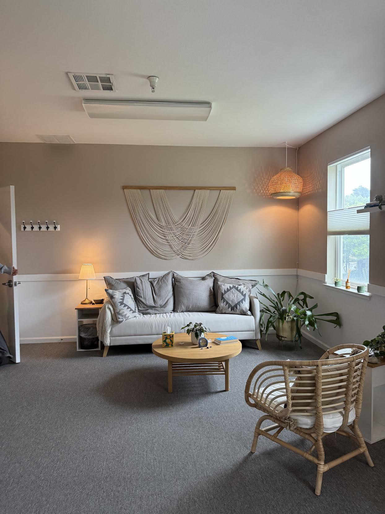 Living room with a white sofa, wooden coffee table, rattan chair, and window with sunlight, beige walls, and decorative wall hanging.