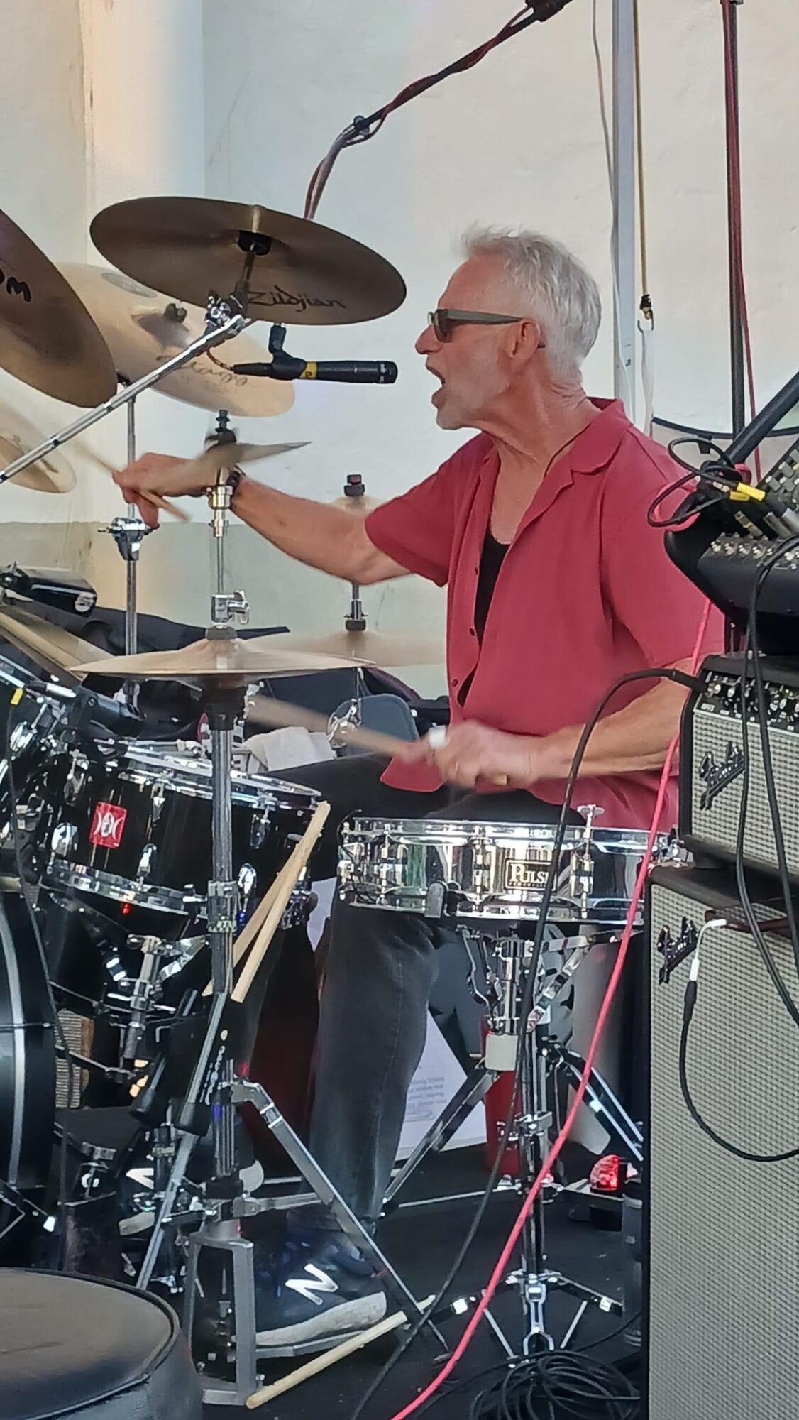 Older man playing drums on stage, wearing glasses and a pink shirt, with musical equipment around him.