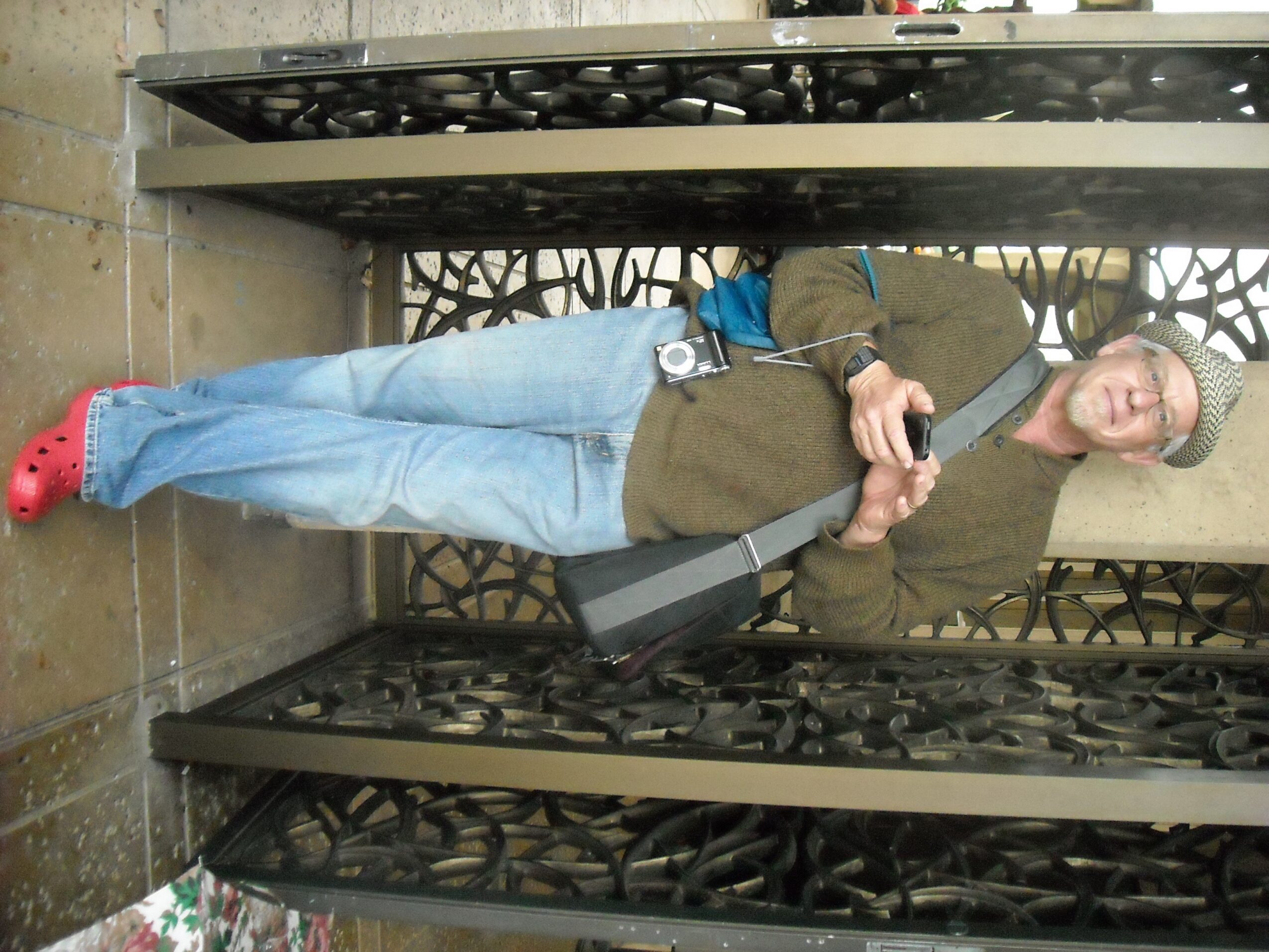 Person standing in an elevator, holding a camera, wearing a green jacket, blue jeans, and red shoes, with a gray bag.