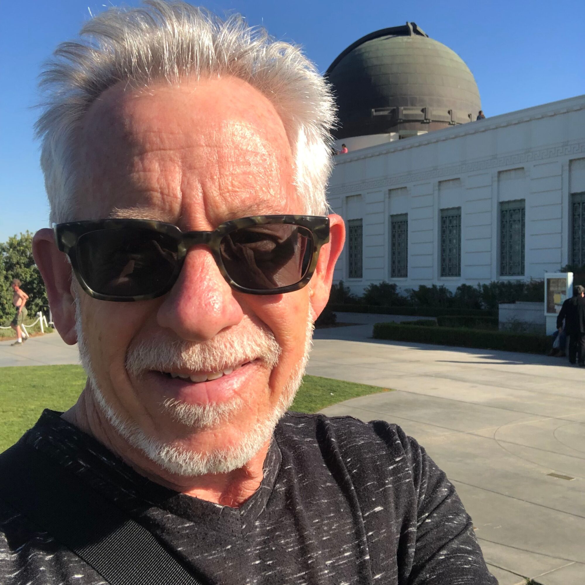 Older man with white hair and beard wearing sunglasses outdoors in front of a white building with columns.