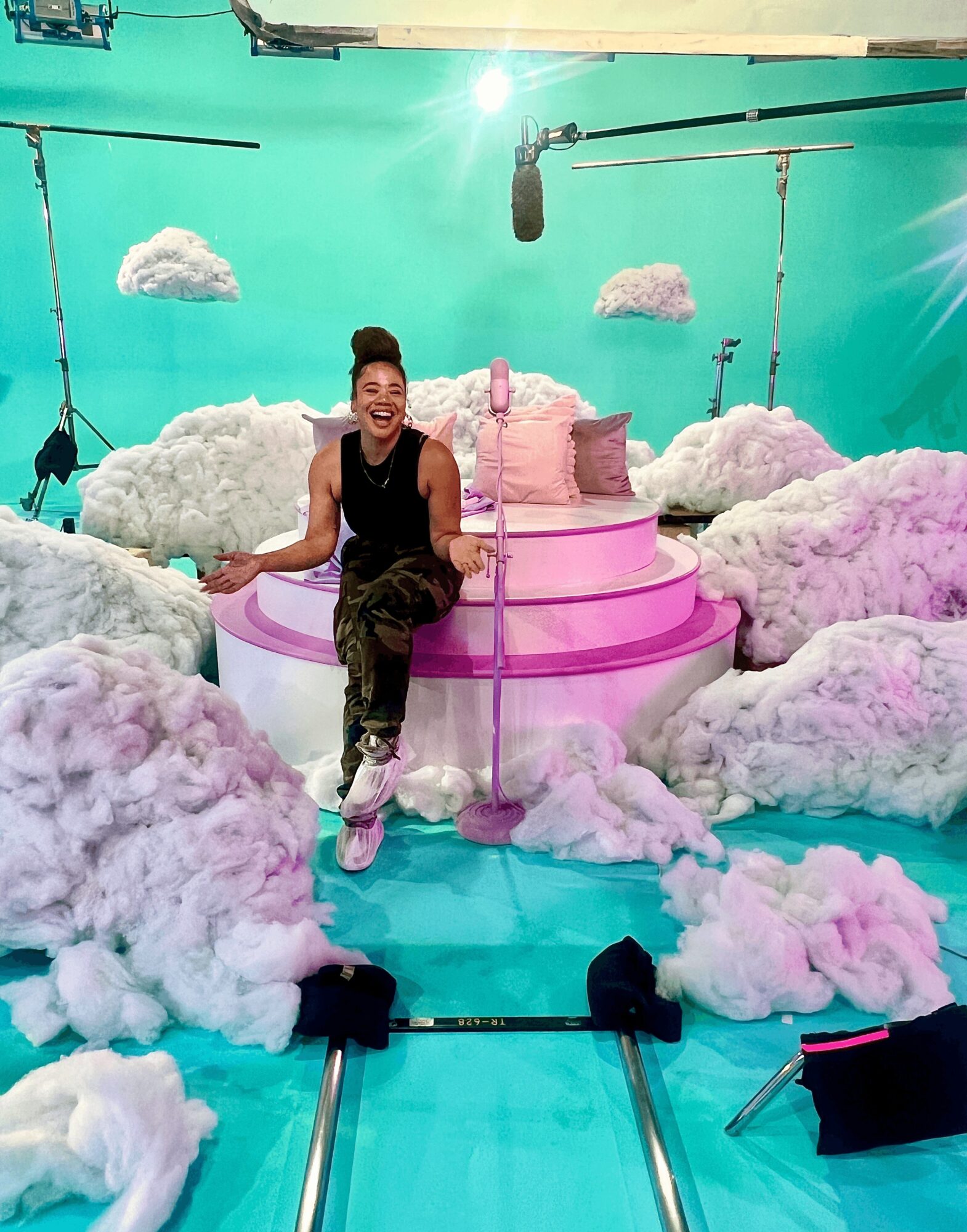 Woman sitting on a pink and white cloud-themed set with clouds and a turquoise background, smiling and holding a pink staff.