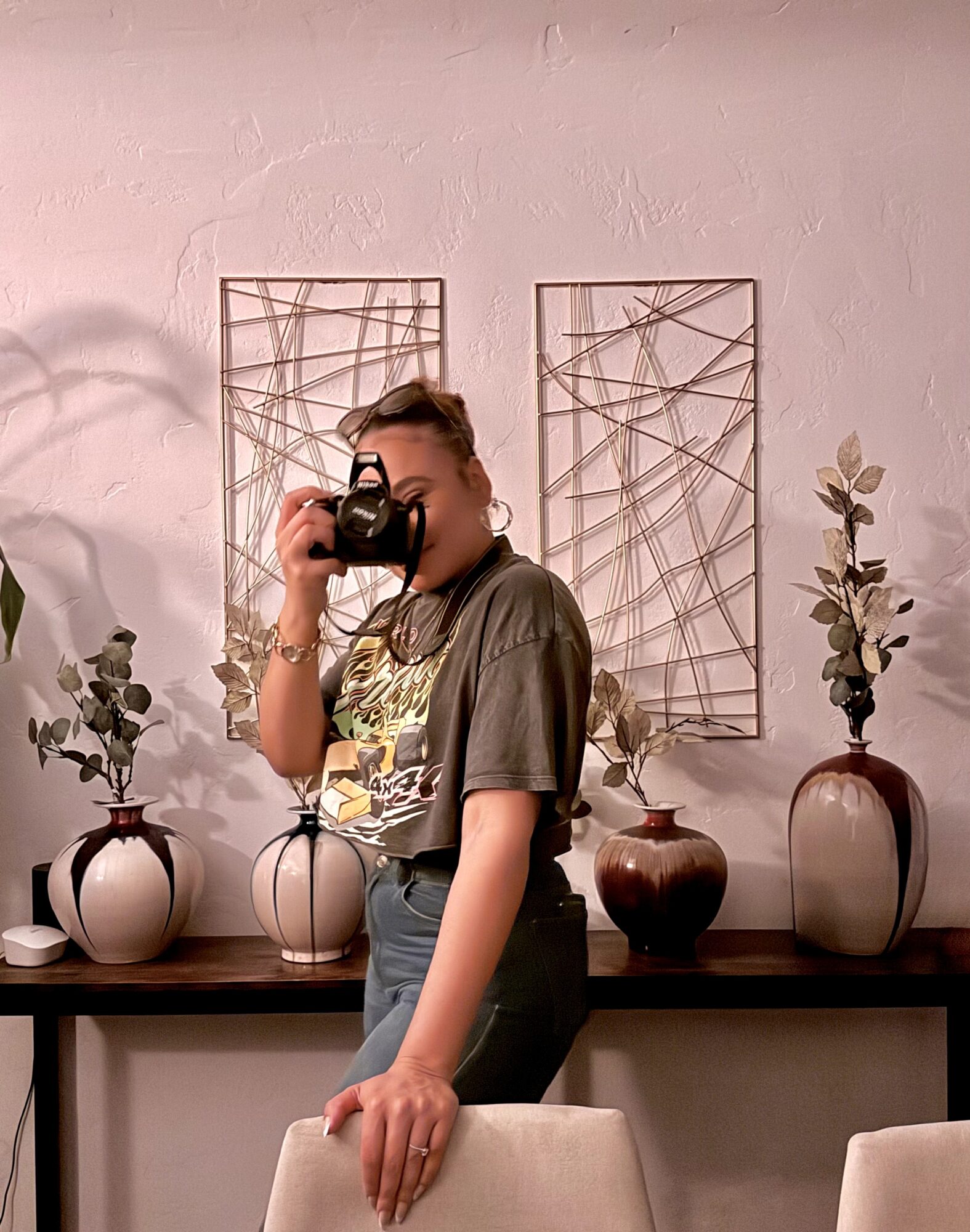 Person taking a photo with a camera, standing in front of a wall with decorative art and vases with plants.