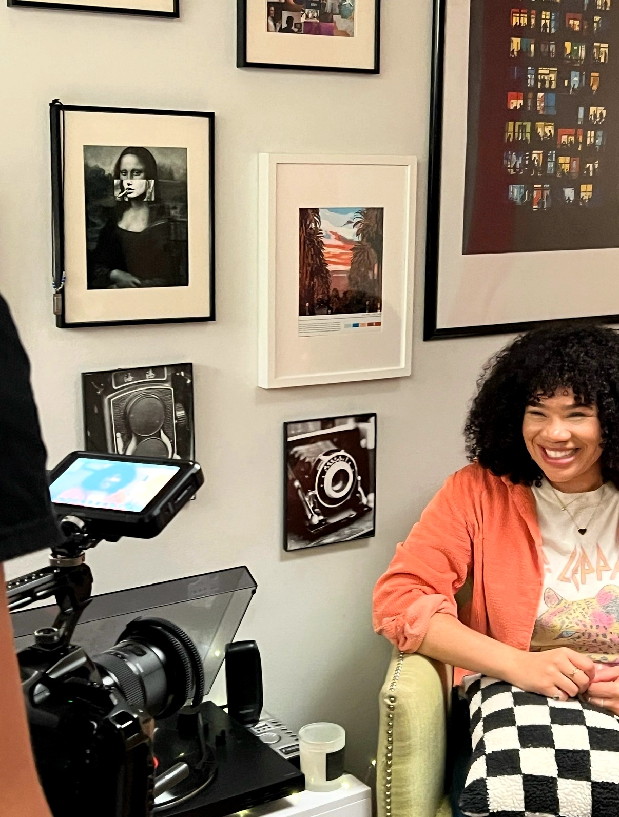 A woman with curly hair smiling, sitting on a green chair, wearing an orange jacket and checkered skirt, in a gallery with framed photos on the wall.