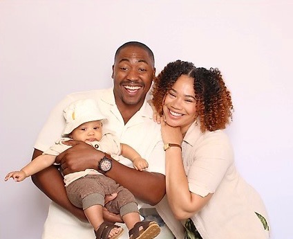 A smiling man holding a baby, with a woman smiling beside them, against a plain background.