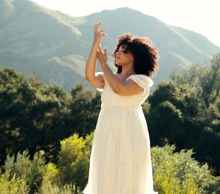 Woman in white dress standing outdoors with mountains and trees in background, raising her hands gracefully.