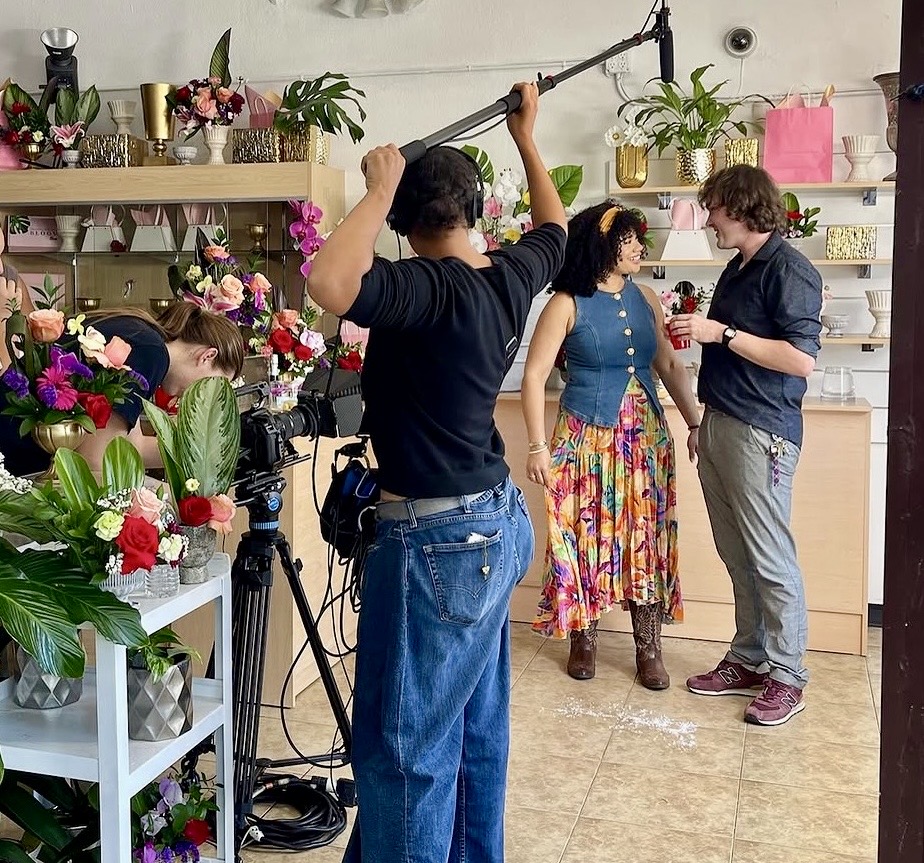 Filming a woman in a floral skirt and blue top with a man in a dark shirt inside a flower shop.