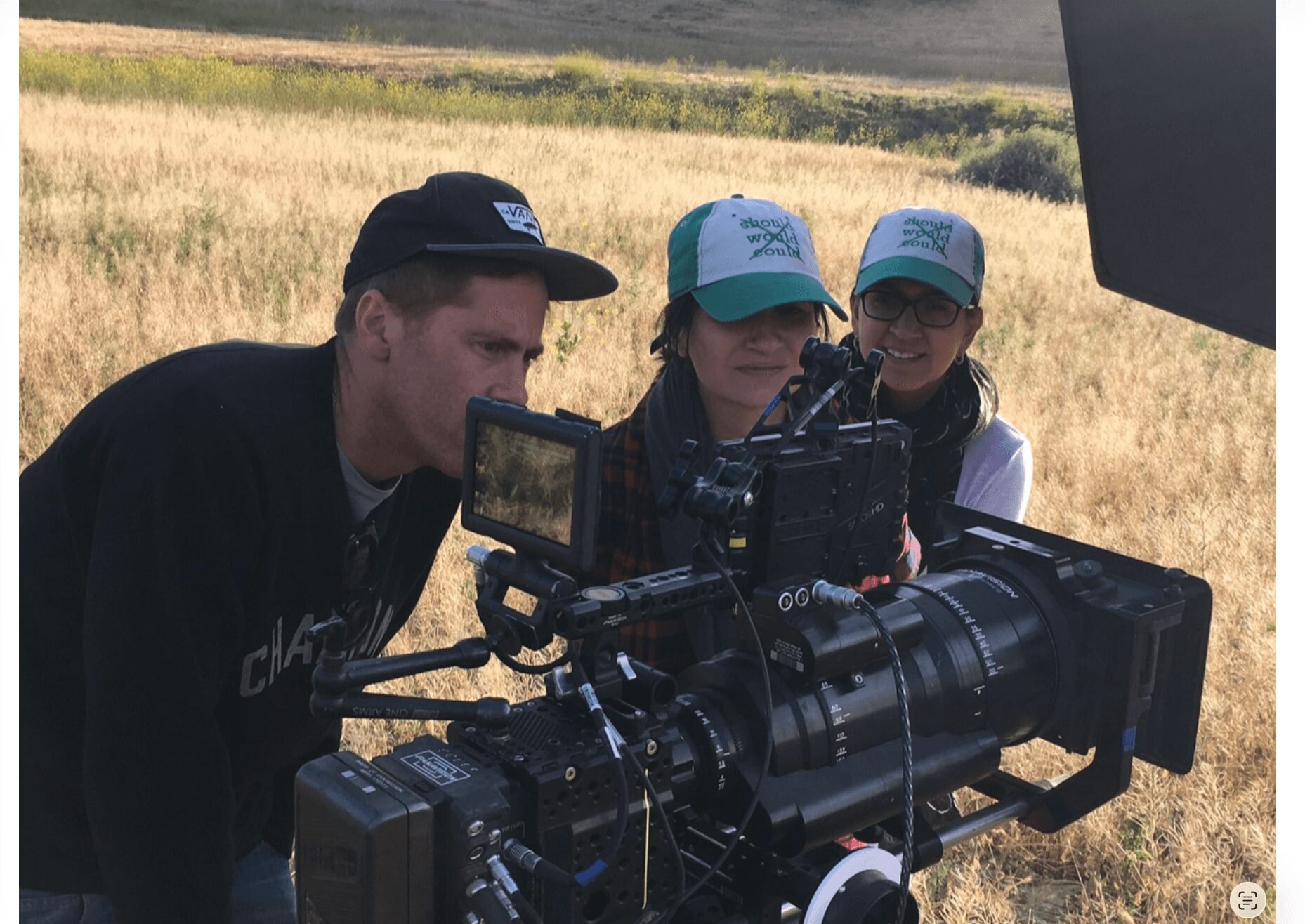 Three people, two children and one adult, look at a large camera or telescope outdoors in a grassy field.