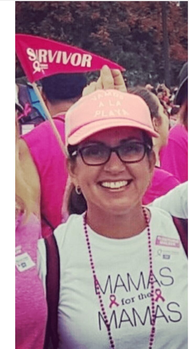 Smiling woman wearing glasses and a pink cap at a breast cancer awareness event, with a pink survivor flag in background.
