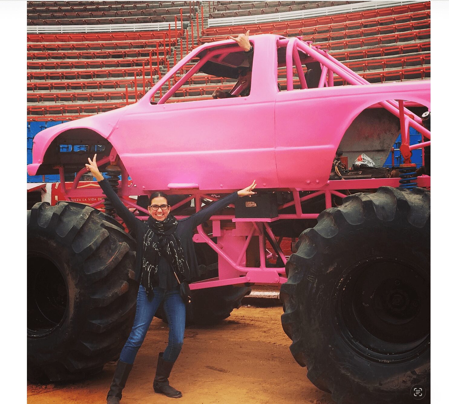 Person standing in front of a large pink monster truck inside a stadium, with arms raised and a smile.
