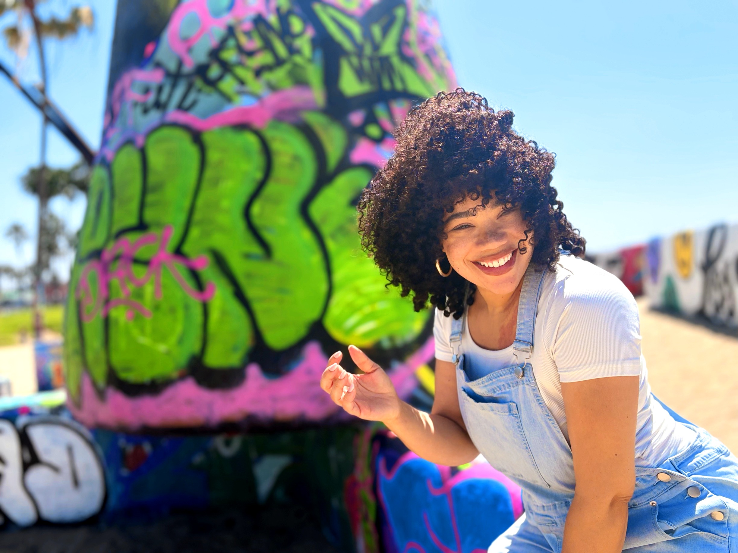 Smiling woman with curly hair in a white shirt and overalls outdoors, graffiti-covered background, sunny day.