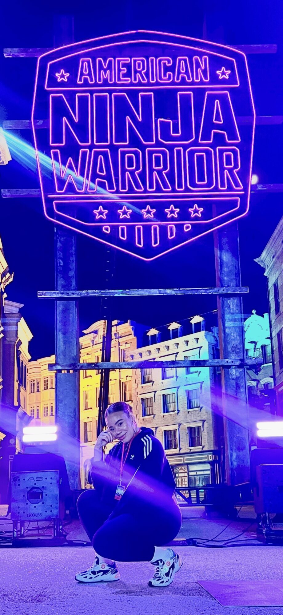 Person crouching in front of a neon sign that reads 'American Ninja Warrior' with city buildings in background.