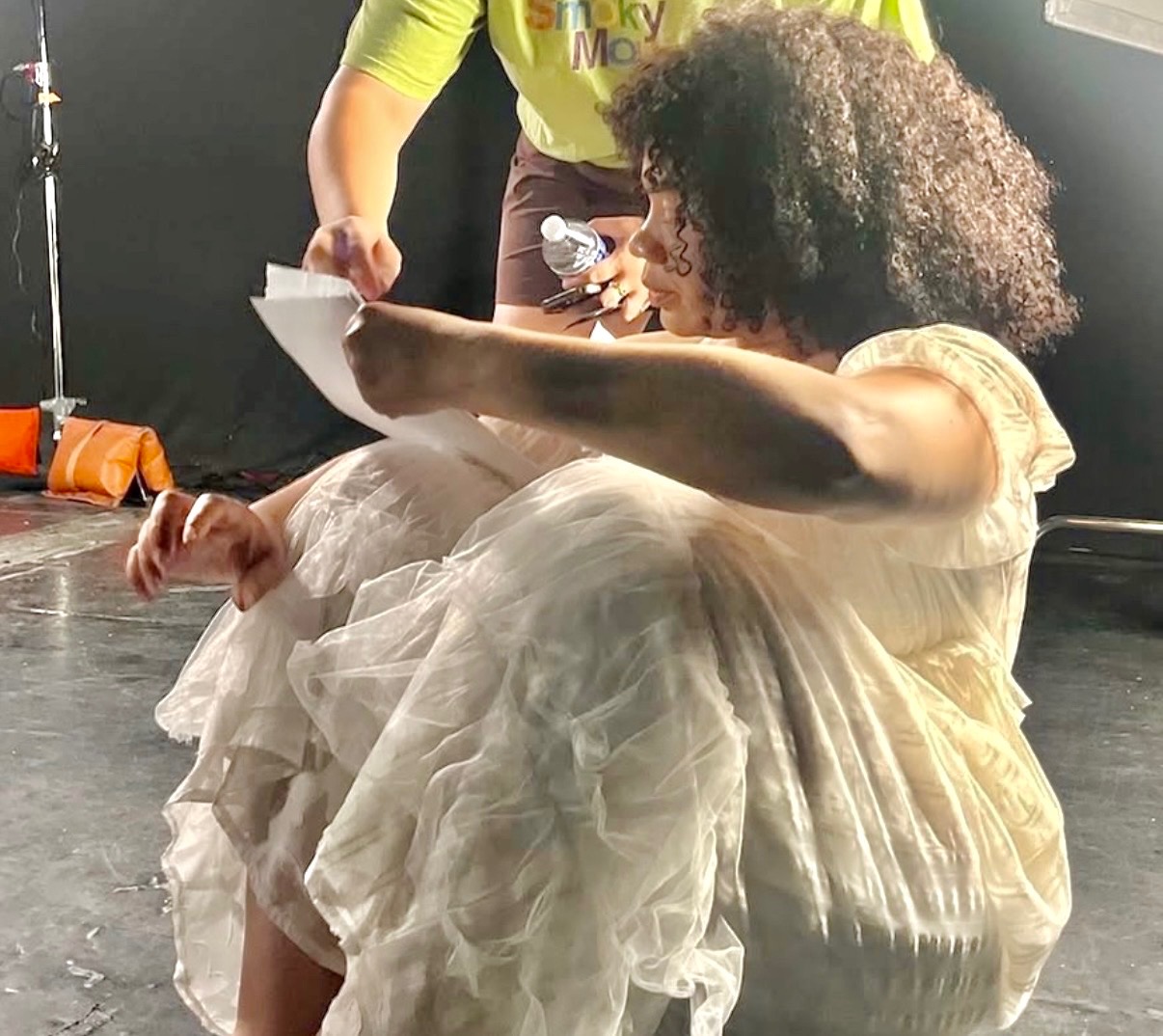 Woman with curly hair sitting on the floor in a light-colored dress, with a person standing nearby holding papers, in a studio setting.