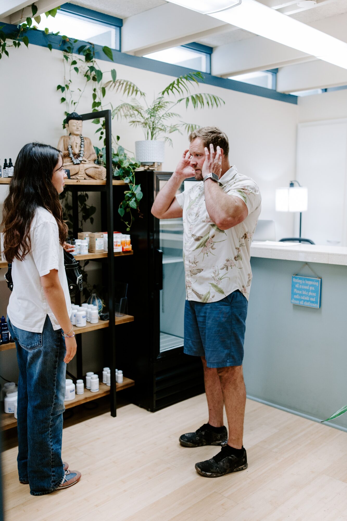 Two people in a room with plants and shelves, one covering ears, the other facing them, in a casual setting.