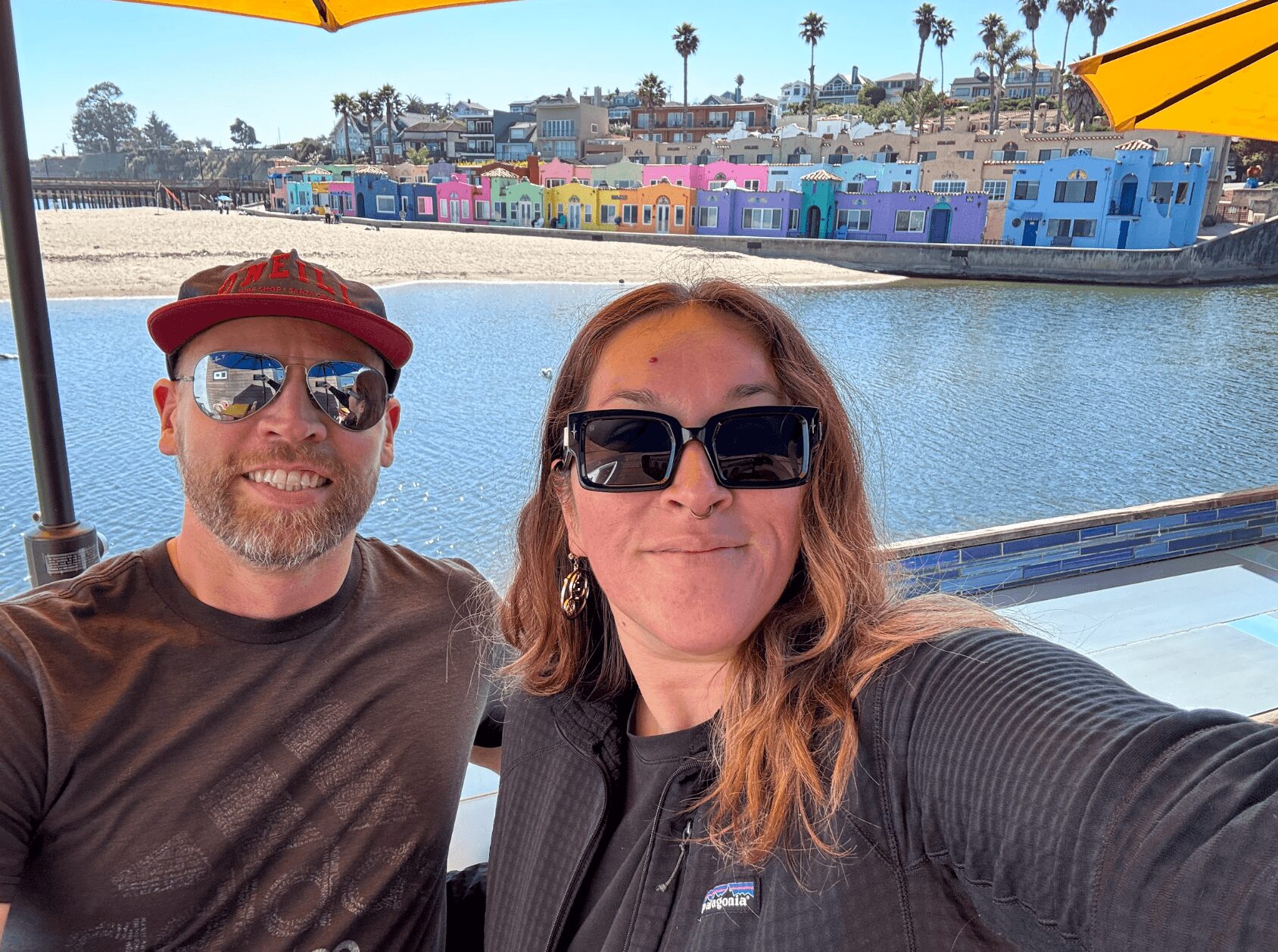Two people taking a selfie outdoors near water with colorful buildings in the background, under yellow umbrellas.