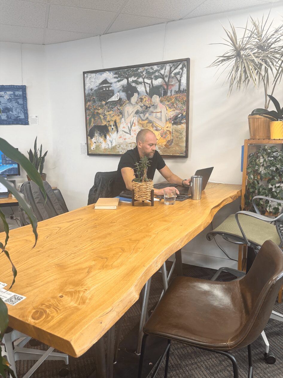 Man working on laptop at a large wooden table in a room with artwork and plants.