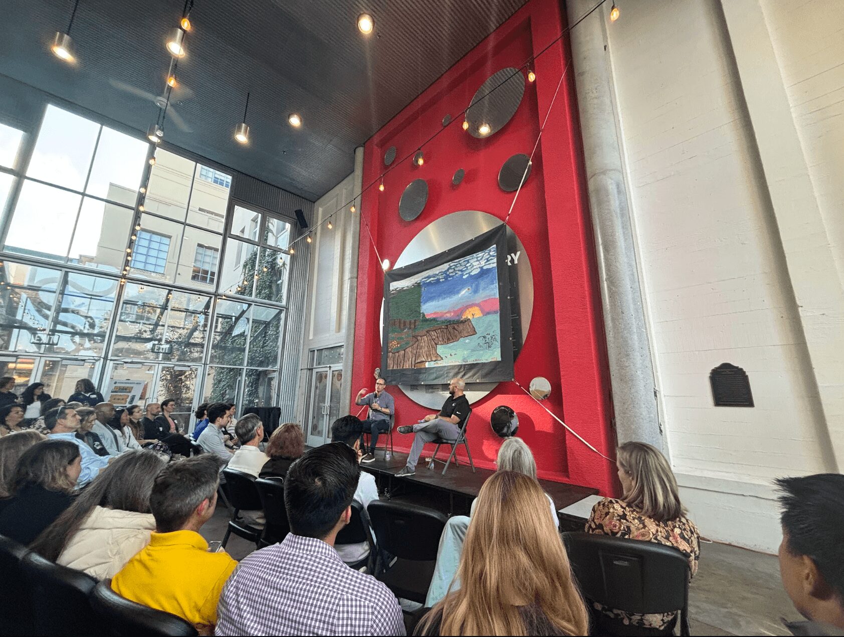 People seated in a modern conference room with large windows, a red wall with a screen, and a speaker presenting.