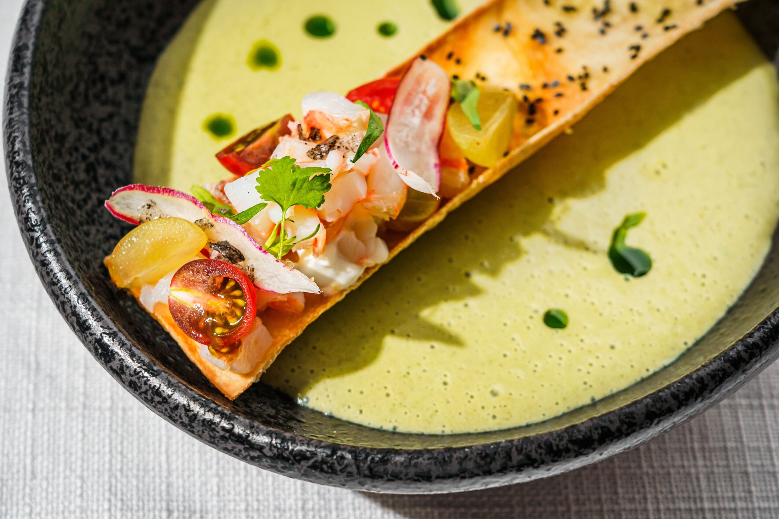 A dish with a rolled wrap filled with vegetables and seafood, served with green sauce in a black bowl.