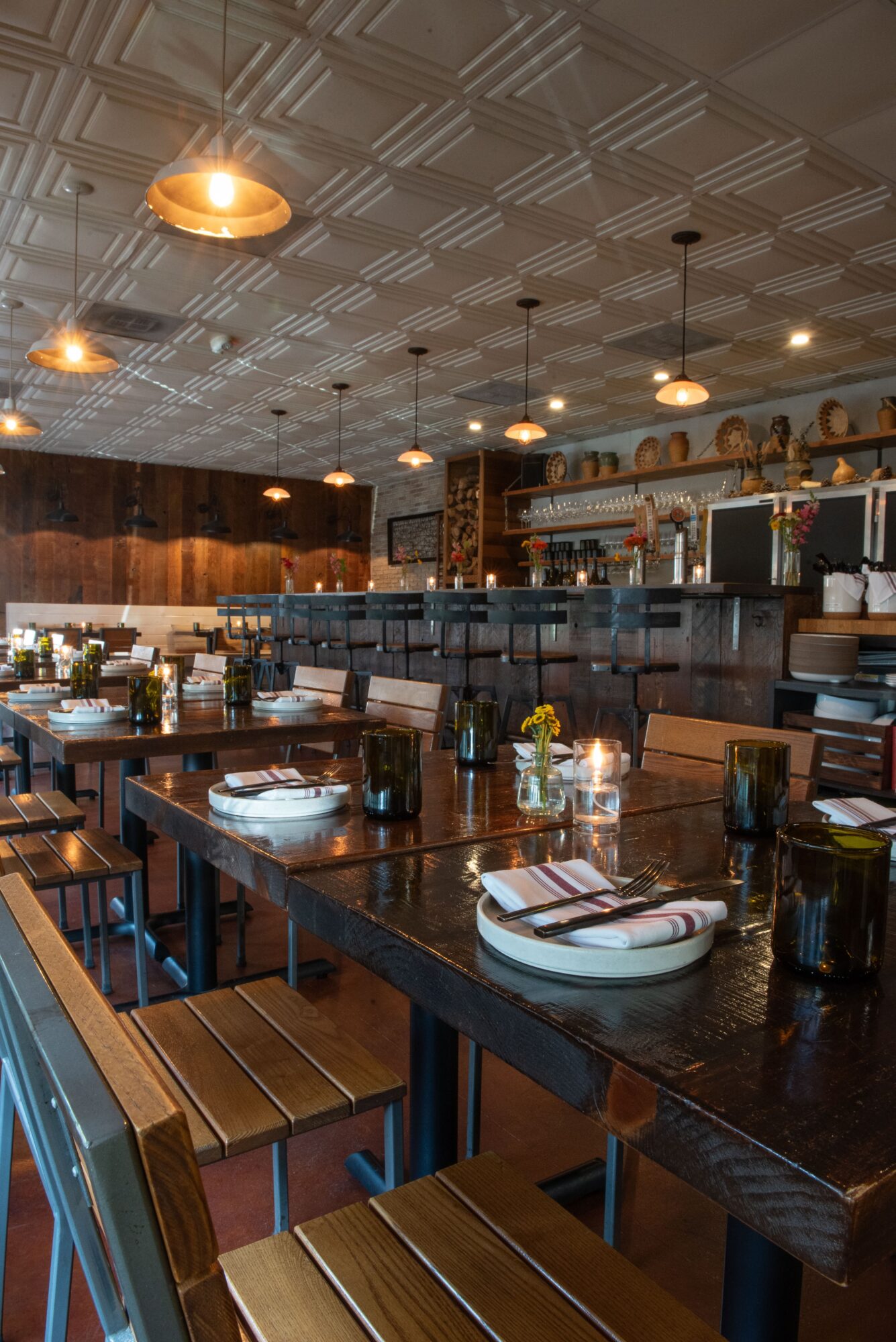 Interior of a restaurant with wooden tables, chairs, pendant lights, and a bar area in the background.