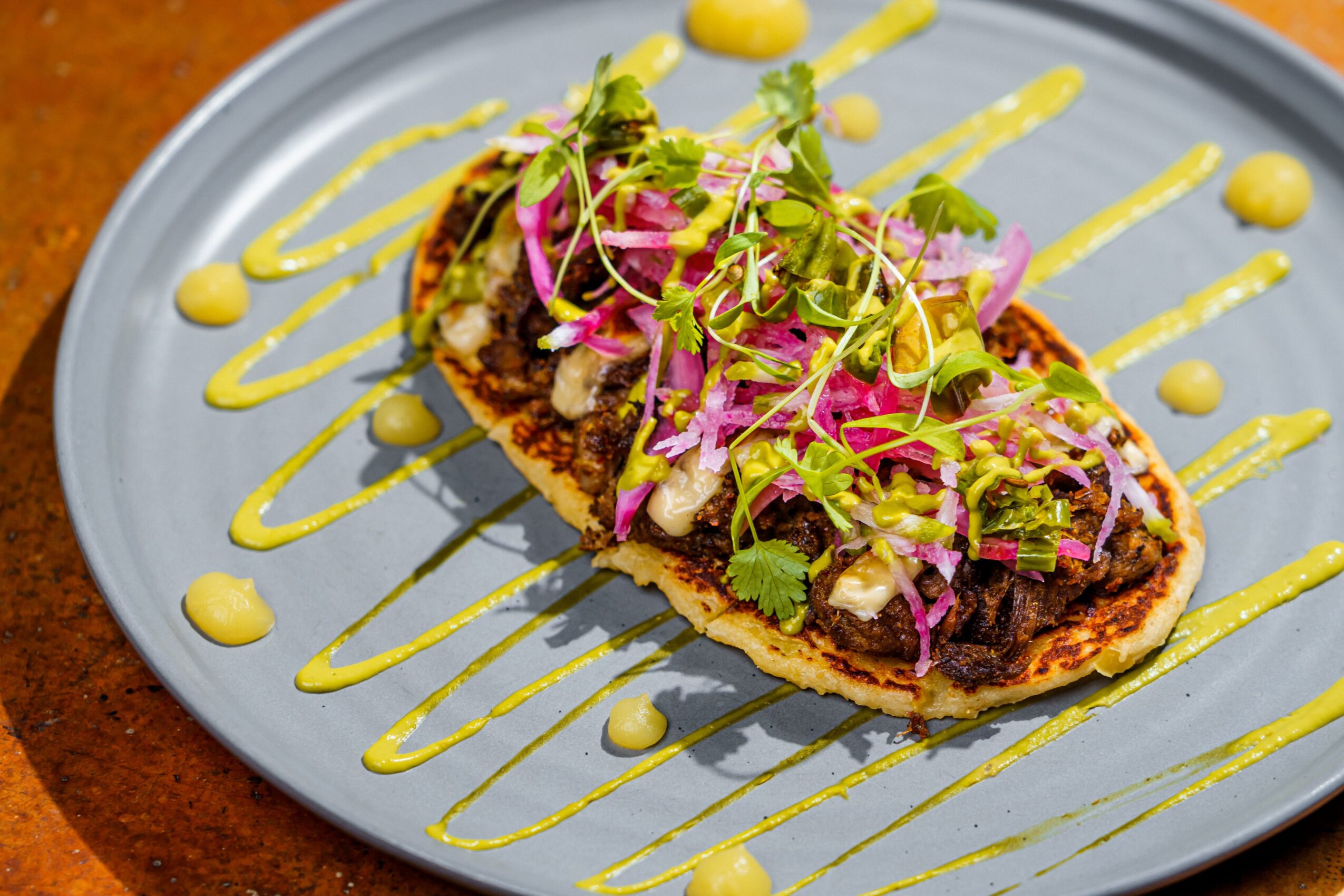 A dish with a stuffed, breaded item topped with microgreens and pink onion slices, drizzled with yellow sauce on a blue plate.